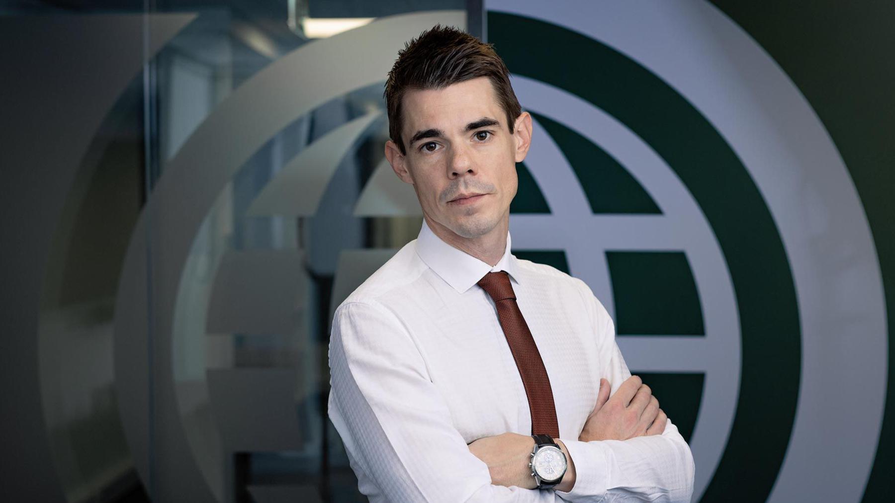 Dan Sexton standing in front of a company logo. He is looking at the camera and is wearing a white shirt and a burgundy tie. He has short, dark hair