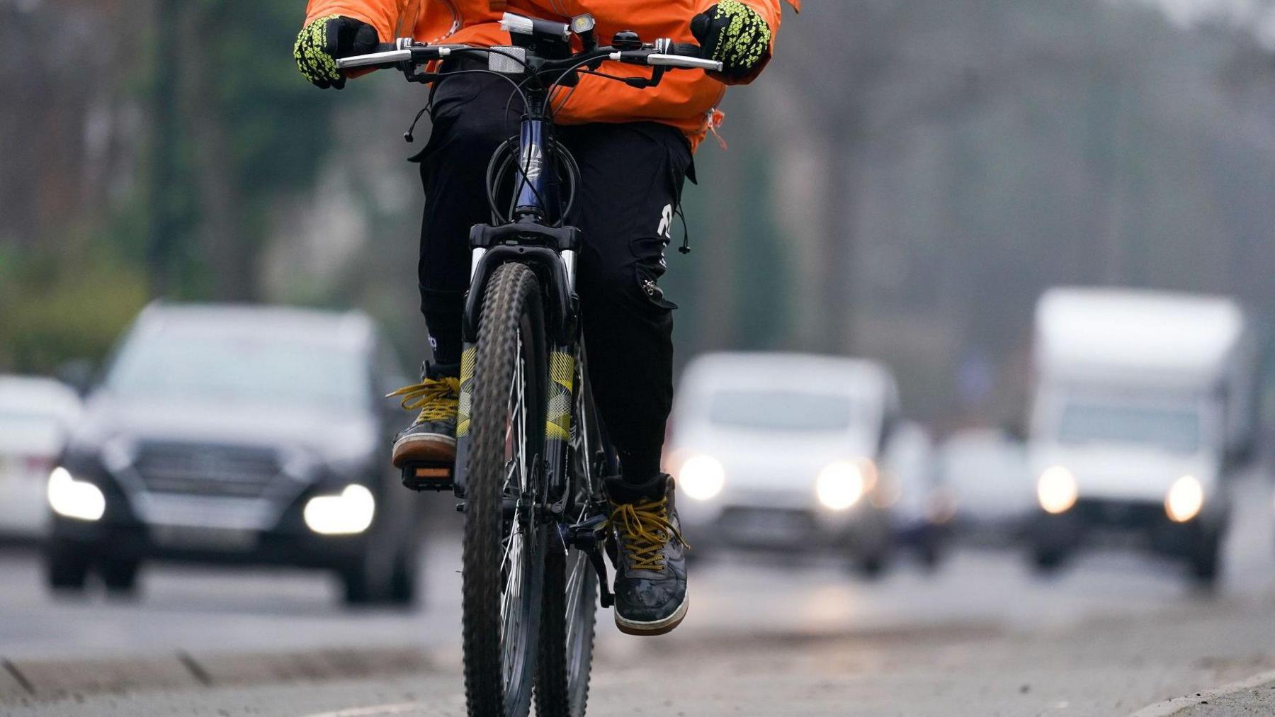 The legs of a cyclist riding a bike, wearing black and yellow gloves, an orange coat and walking boots. There are pixelated vehicles in the background. 