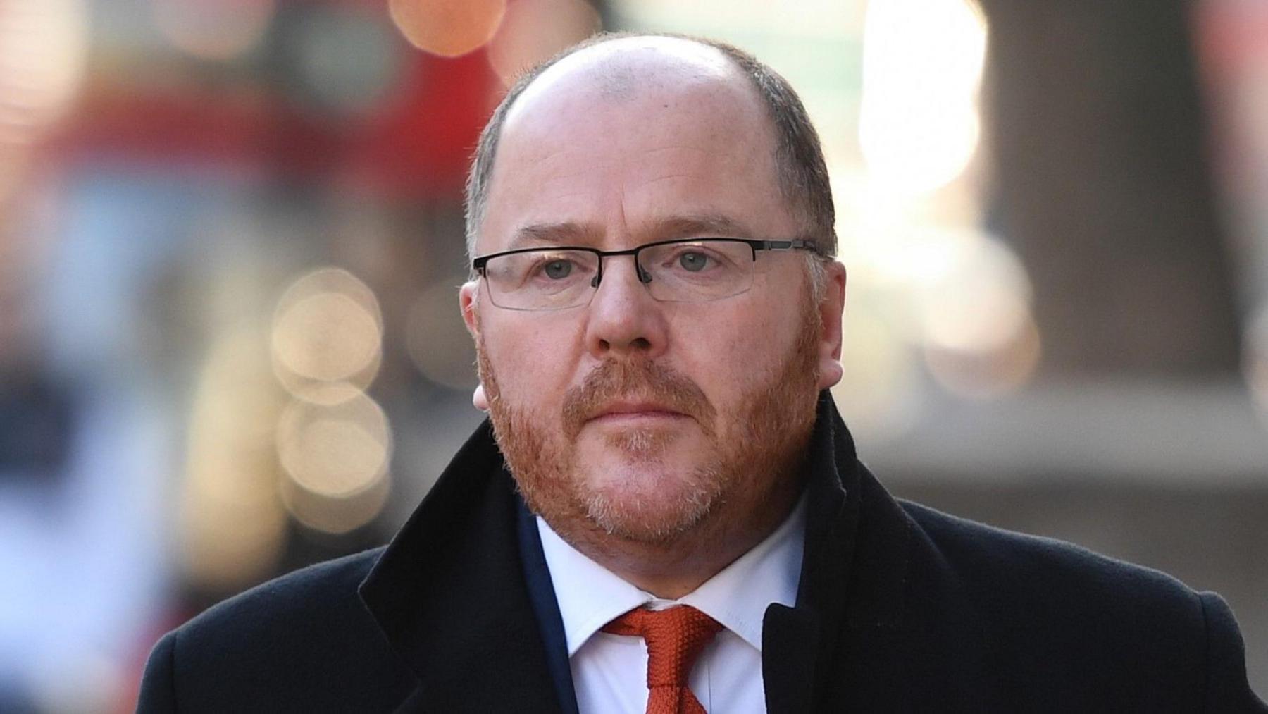 George Freeman, who has very short greying hair and a reddish close-cropped beard. He is wearing glasses and a grey jacket over a white shirt and blue tie.