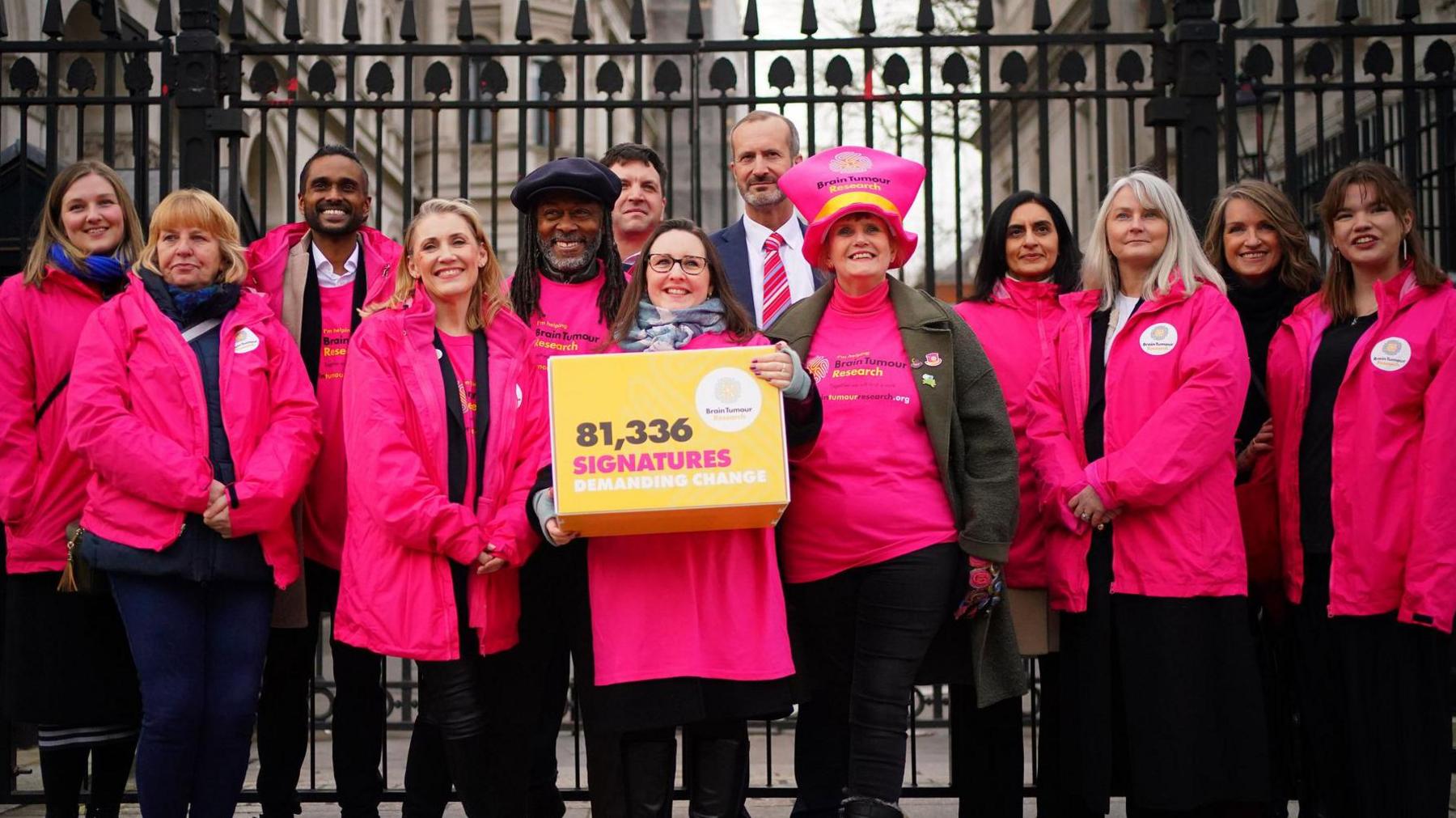 A radical  of 13 radical   basal   by the gates for Downing Street wearing pinkish  jackets and Theo Burrell is successful  the mediate  holding a yellowish  container  that reads "81,336 signatures demanding change"