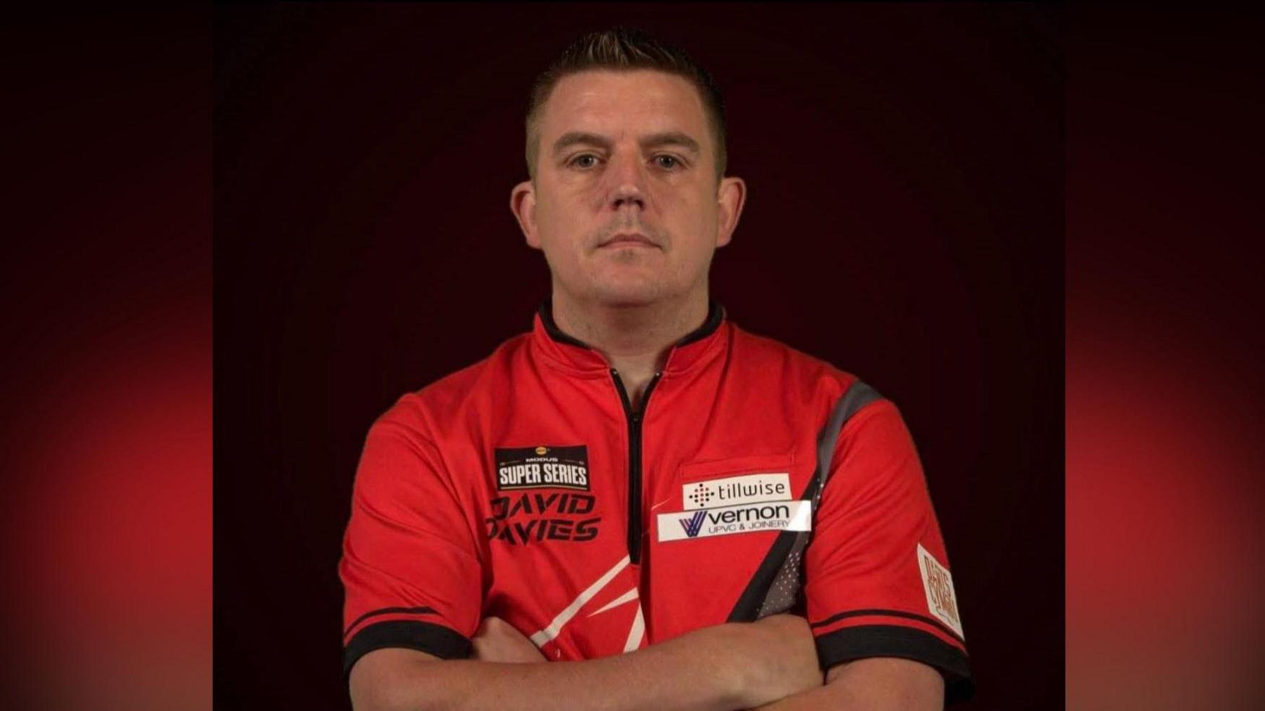 Llun o David Davies yn sefyll gyda'i freichiau wedi croesi o'i flaen wrth edrych at y camera. Mae ganddo wallt brown byr ac mae'n gwisgo crys-t coch