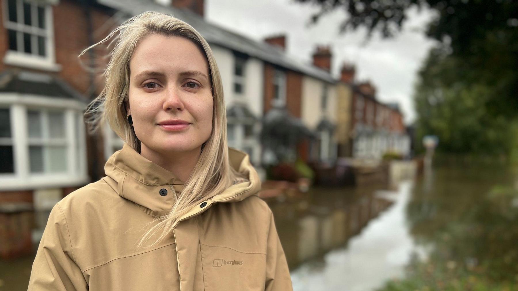 A woman wearing a beige hooded coat standing in front of a row of flooded houses