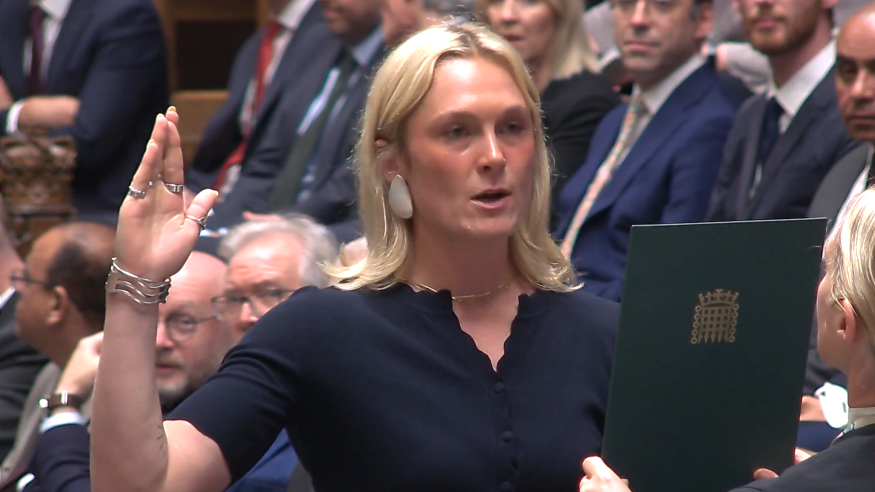 Lara Bird, who has shoulder length blonde hair, stand with her right hand raised as she reads off a green sheet held to her face. 