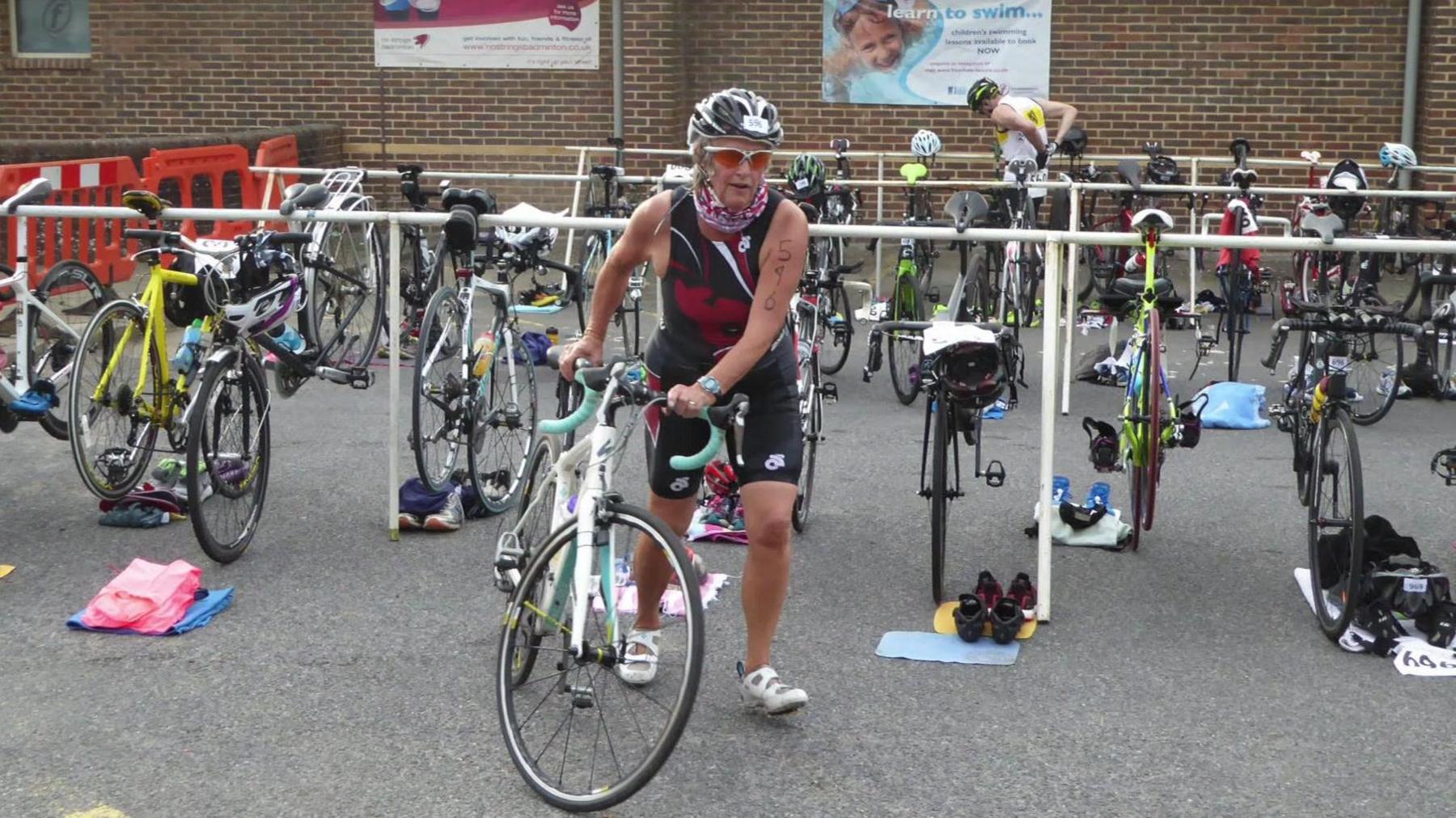 A woman in a bike helmet, sunglasses, and a dark-coloured triathlon kit pushing a bicycle. There are several other bicycles fixed to railings behind her.