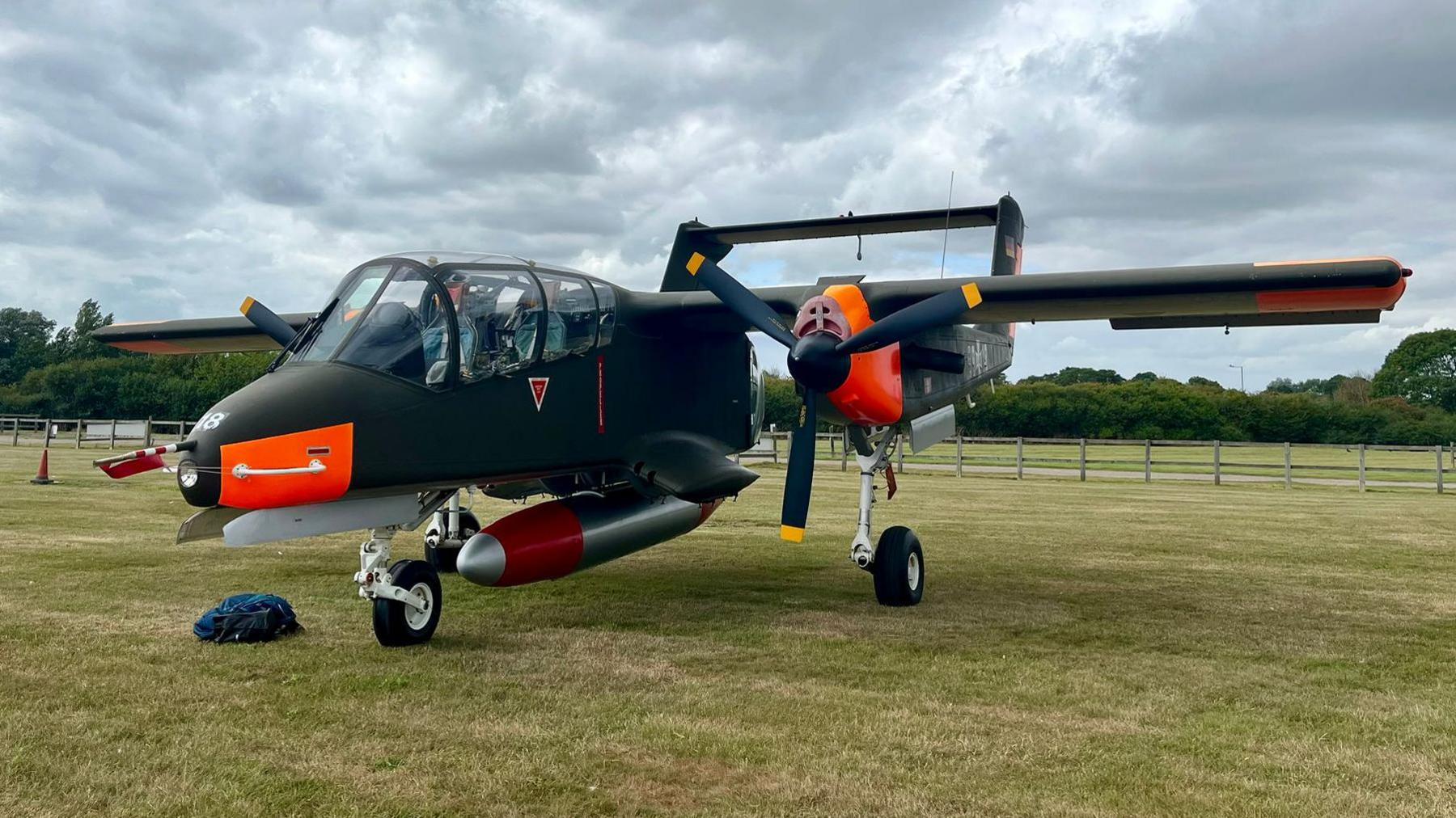 A green Bronco aircraft, which has propellers beneath its wings that are held on by orange fittings. The plane is parked in a large field.