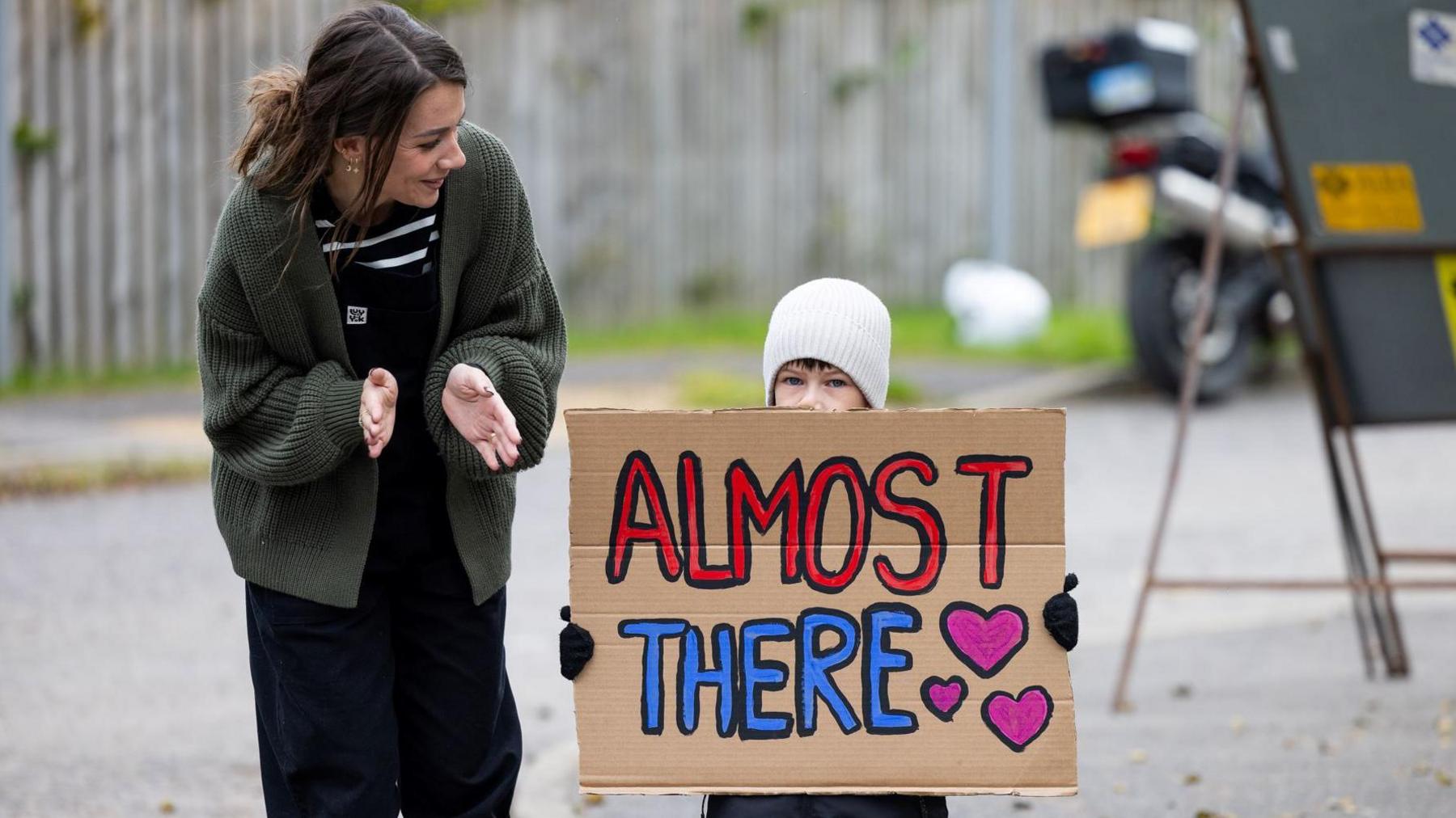 A woman clapping her hands as she stands next to a child holding up a cardboard sign with "Almost there" and pink hearts on it