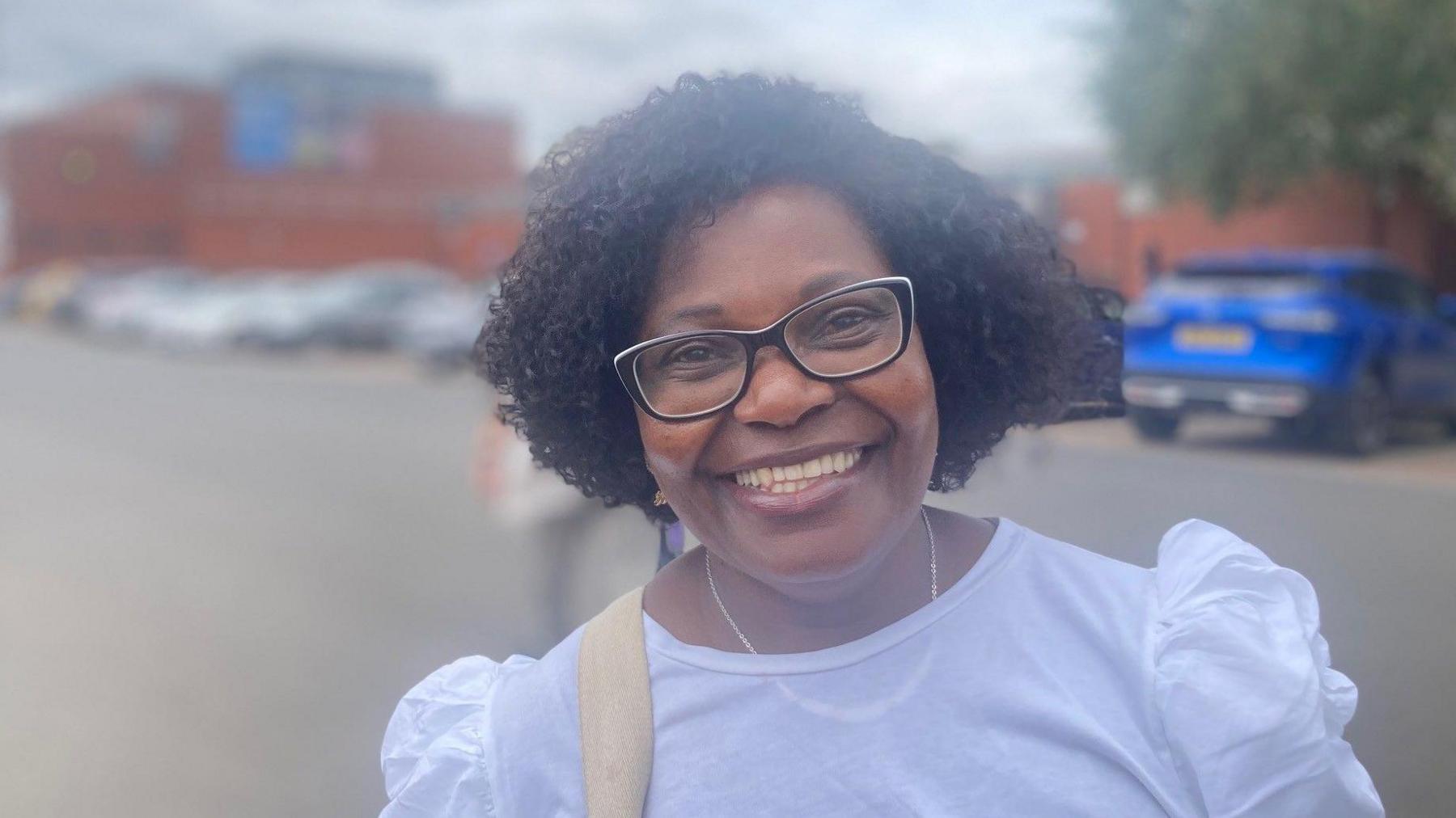 A woman with short curly black hair and glasses smiles at the camera. She is wearing a white blouse and a necklace. Behind her are parked cars, and a red-brick building.