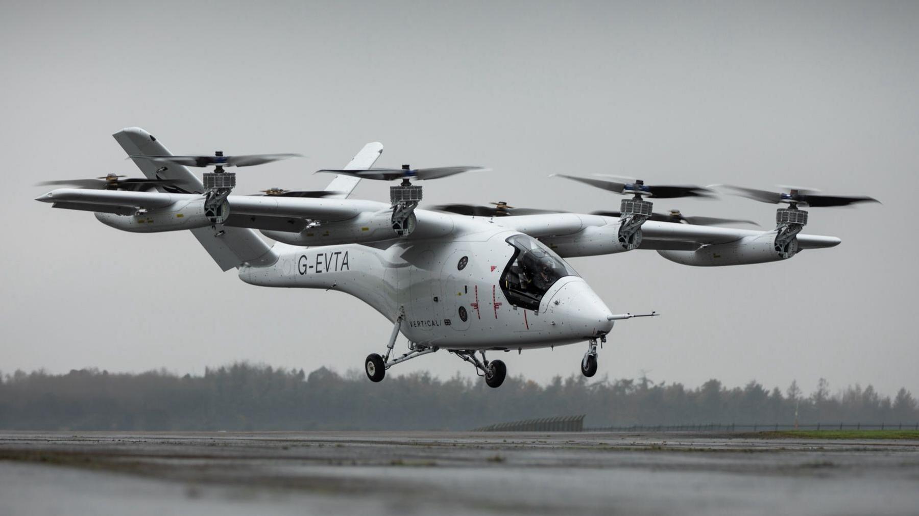 A small aircraft lifts off the runway at Cotswold Airport. There are eight small rotors attached to wings on both sides of the fuselage, and a v-shaped tailfin.