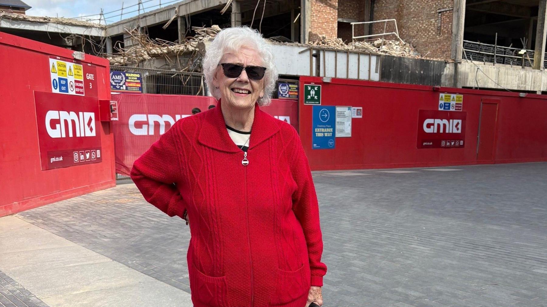 A woman with curly short grey hair wears a grey cardigan. A building site is in the background.