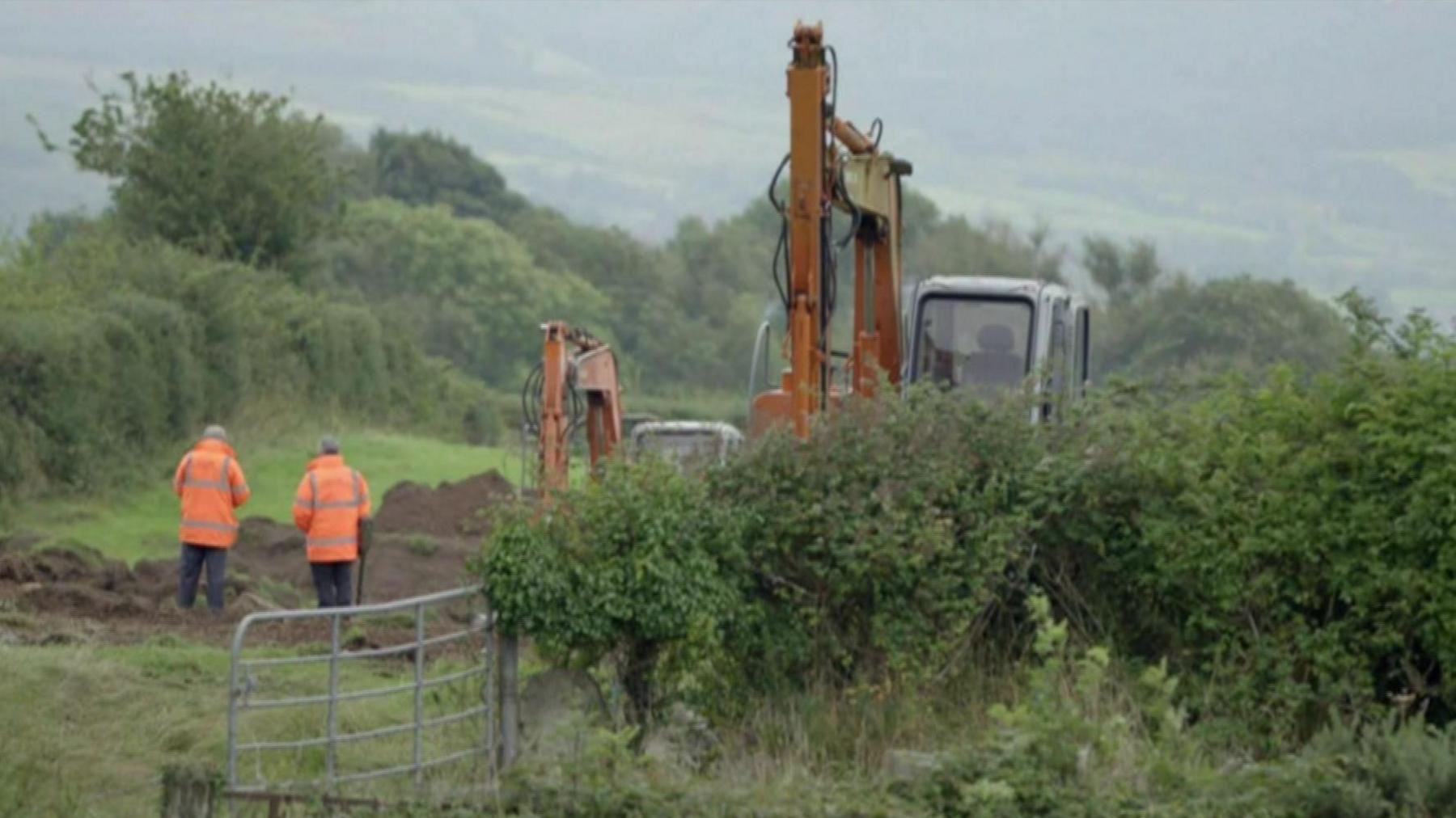 A screengrab of TV footage of the ICLVR's search for Capt Robert Nairac.   Two men dressed in orange high-visibility jackets are standing in a field beside an open gate.  Two mechanical diggers are also in the field and a large section of grass has been dug up and turned over for examination.