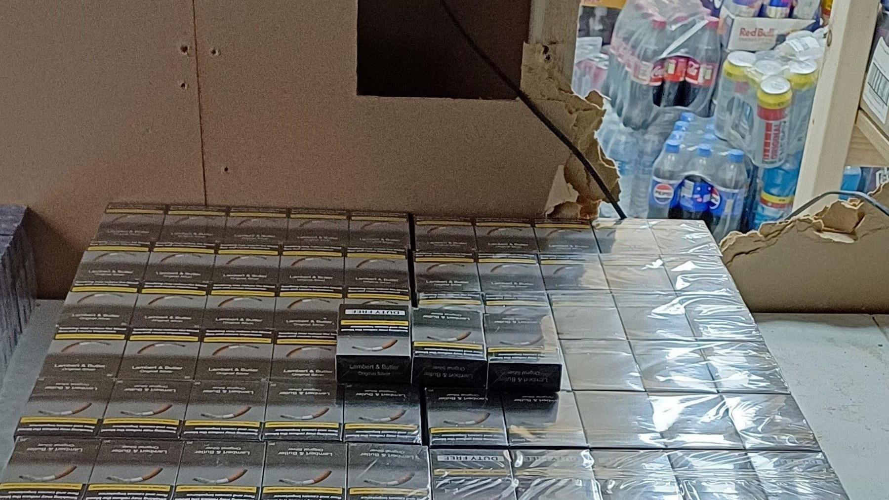 Boxes of cigarettes on a table with a broken wall behind them. Bottles of pop can be seen behind the wall.