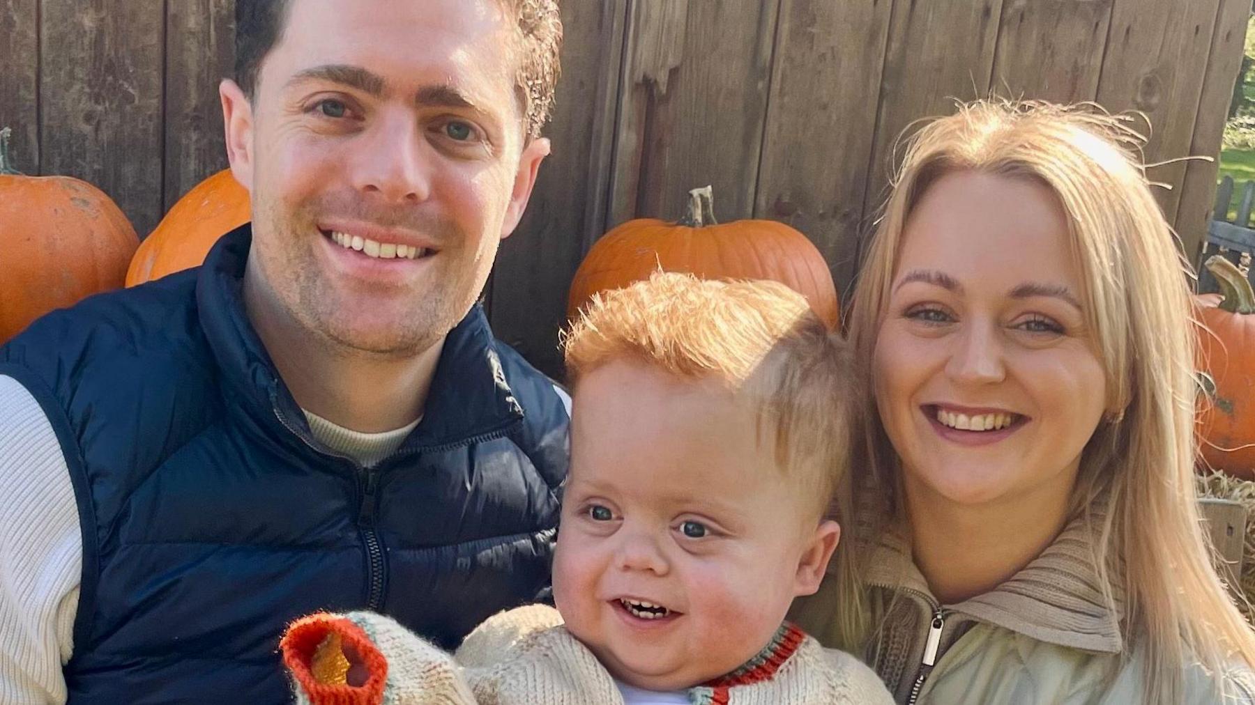 Reece and Shona are smiling at the camera while holding Ari, who is looking to the left of the camera and pointing at something out of shot. They are sat on hay bales with pumpkins behind them.