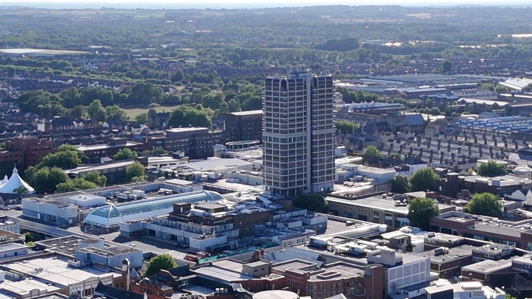 An overhead view of a densely built town centre with a tall tower block in the centre of the frame, as well as the glass roof of a large shopping centre, while greenery stretches into the distance.