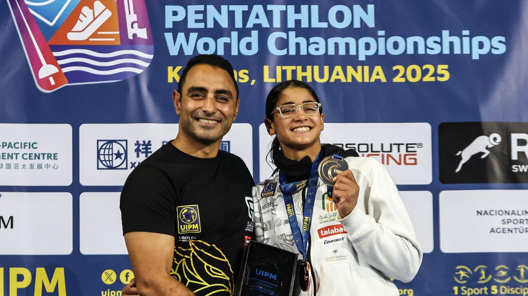 Mohamed Abu Hashem, wearing a black T-shirt with yellow detail on it, stands beside Farida Khalil, who is wearing clear-framed glasses and a white tracksuit top with sponsor badges on the front of it. Both are visible from waist up and are smiling as Khalil holds up a gold medal and Abu Hashem holds a black plaque. They stand on the top step of a podium, which is out of shot, with a sponsor board at the Pentathlon World Championships behind them