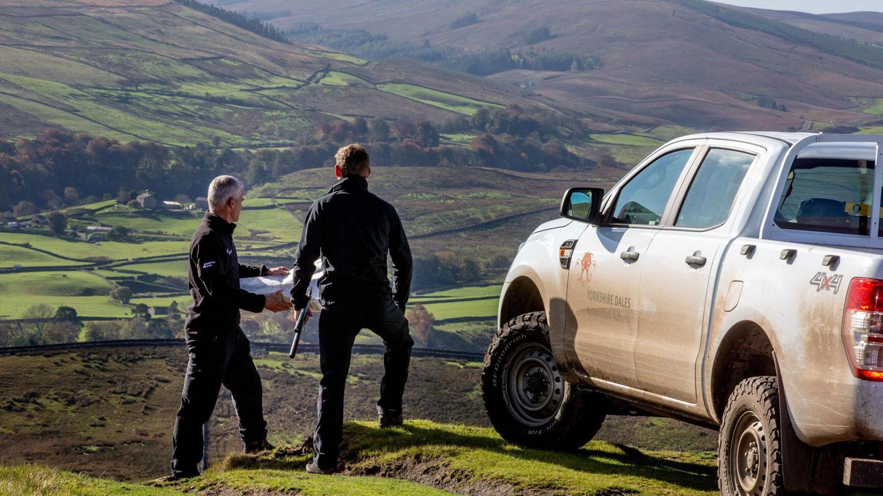 Two men hold paper plans while overlooking moors standing left of a parked up white, mud-splattered 4x4