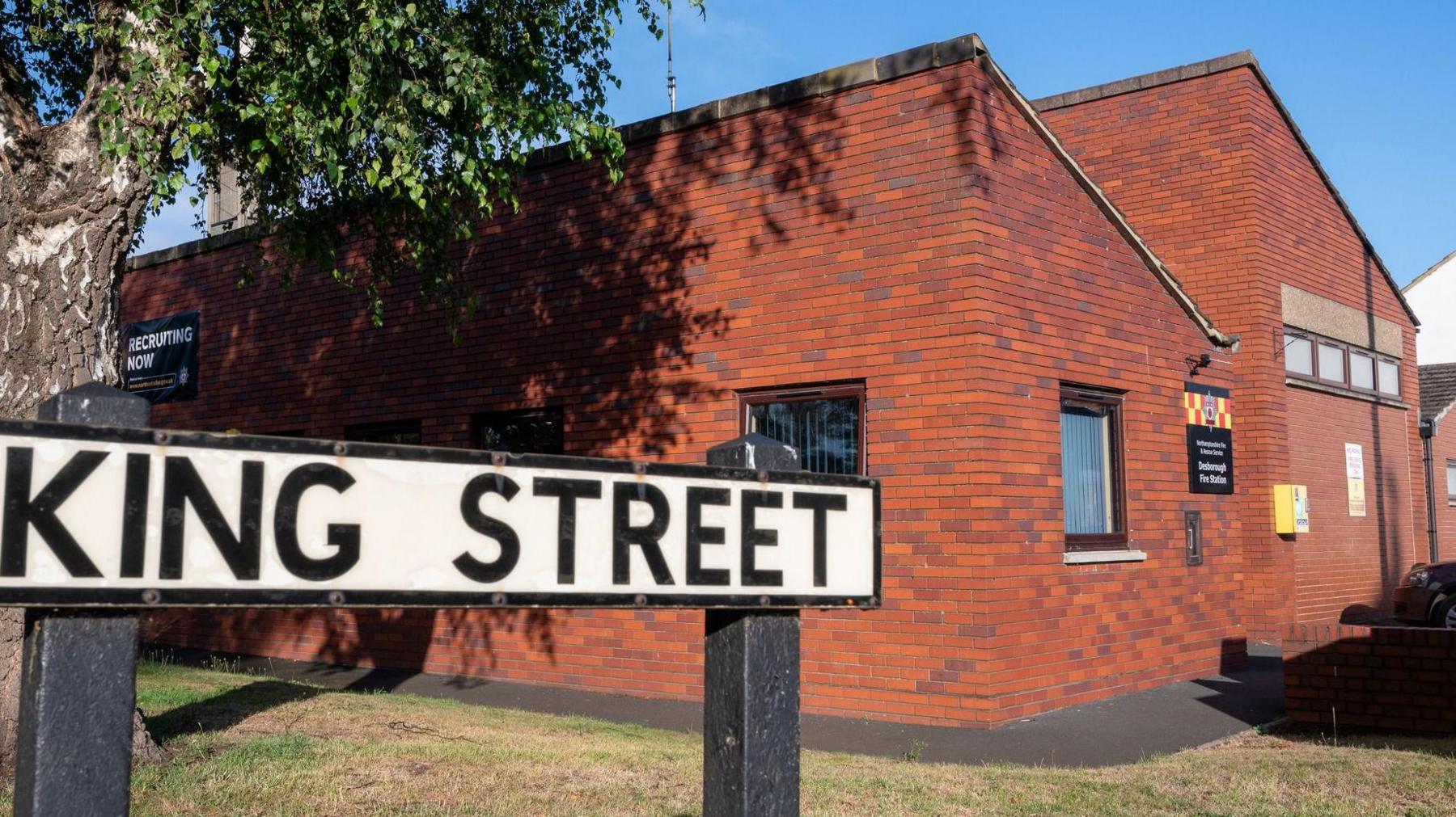 A red brick fire station, built in 1975, with two slanted roofs. There is a street sign in front of the building, saying King Street. There is grass in front and a tree to the left. The building has several small windows. 