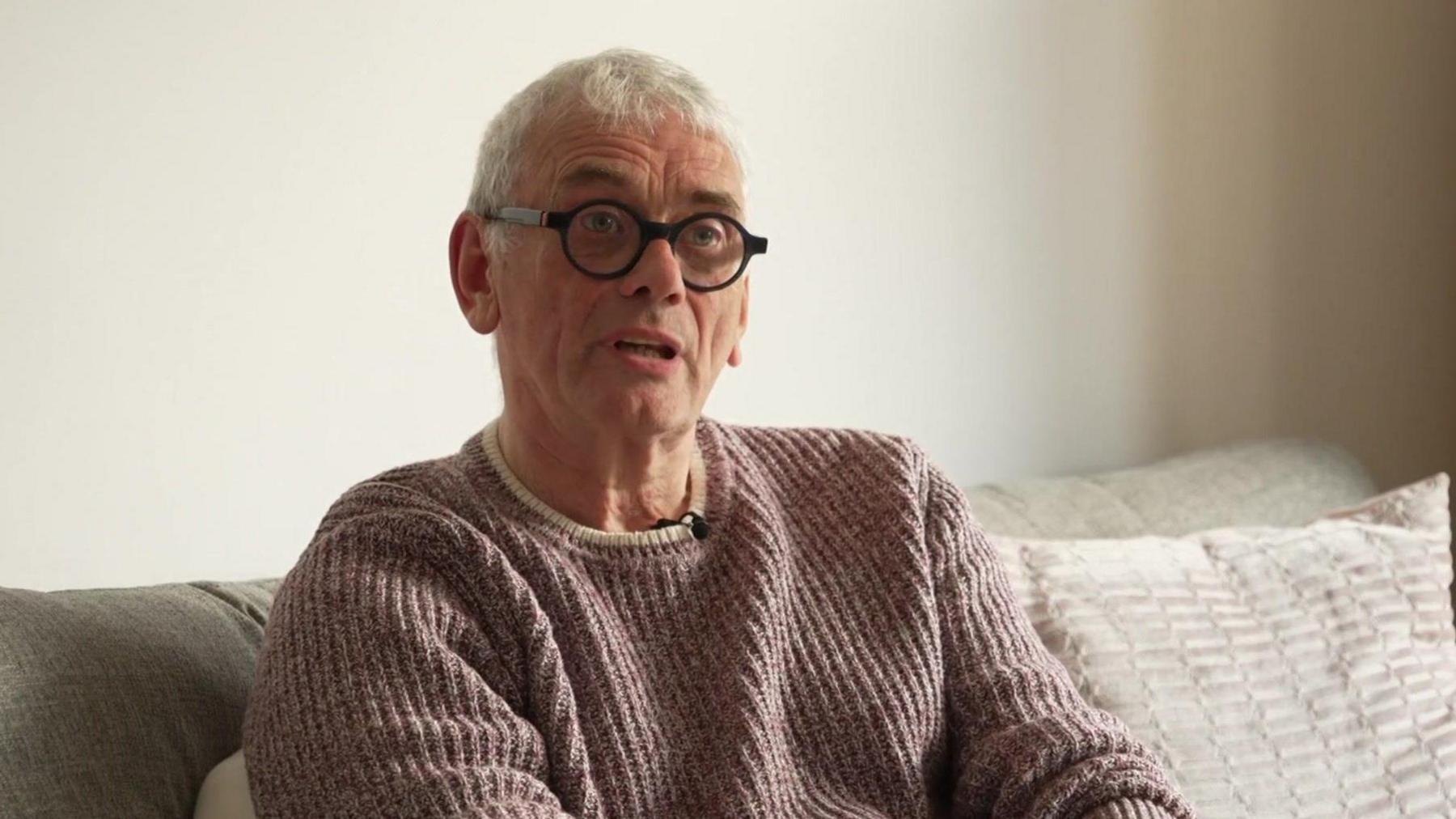 Robin, who has a mauve cable knit jumper, white hair and round black rimmed glasses, speaks to the camera sitting on a grey couch