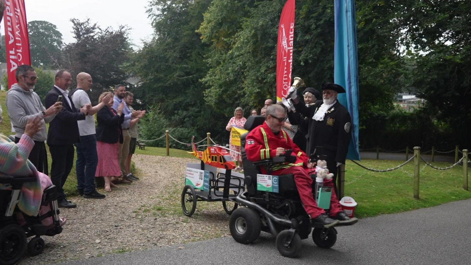 Steven Webb sets off in his wheelchair towing a small model of Cornwall's air ambulance on a trailer behind him as well-wishers applaud and the town crier, standing next to the mayor, rings a bell. 