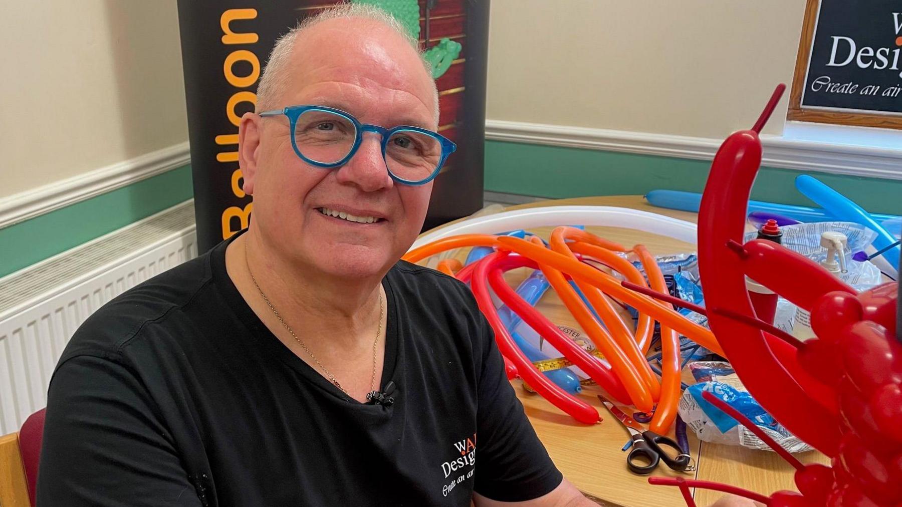 A man in his sixties smiles at the camera. He is sitting next to a table which is covered in modelling balloons and other equipment, including scissors. There are several red balloons in the foreground, which make up the back of one of his dresses. He is wearing bright blue glasses.