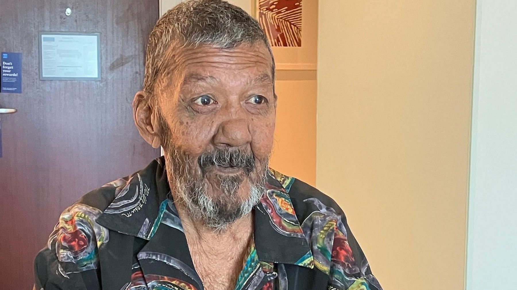 Nizam Hosein with short greying hair and bread looking off camera. He is wearing a black shirt with a mythical, colourful pattern