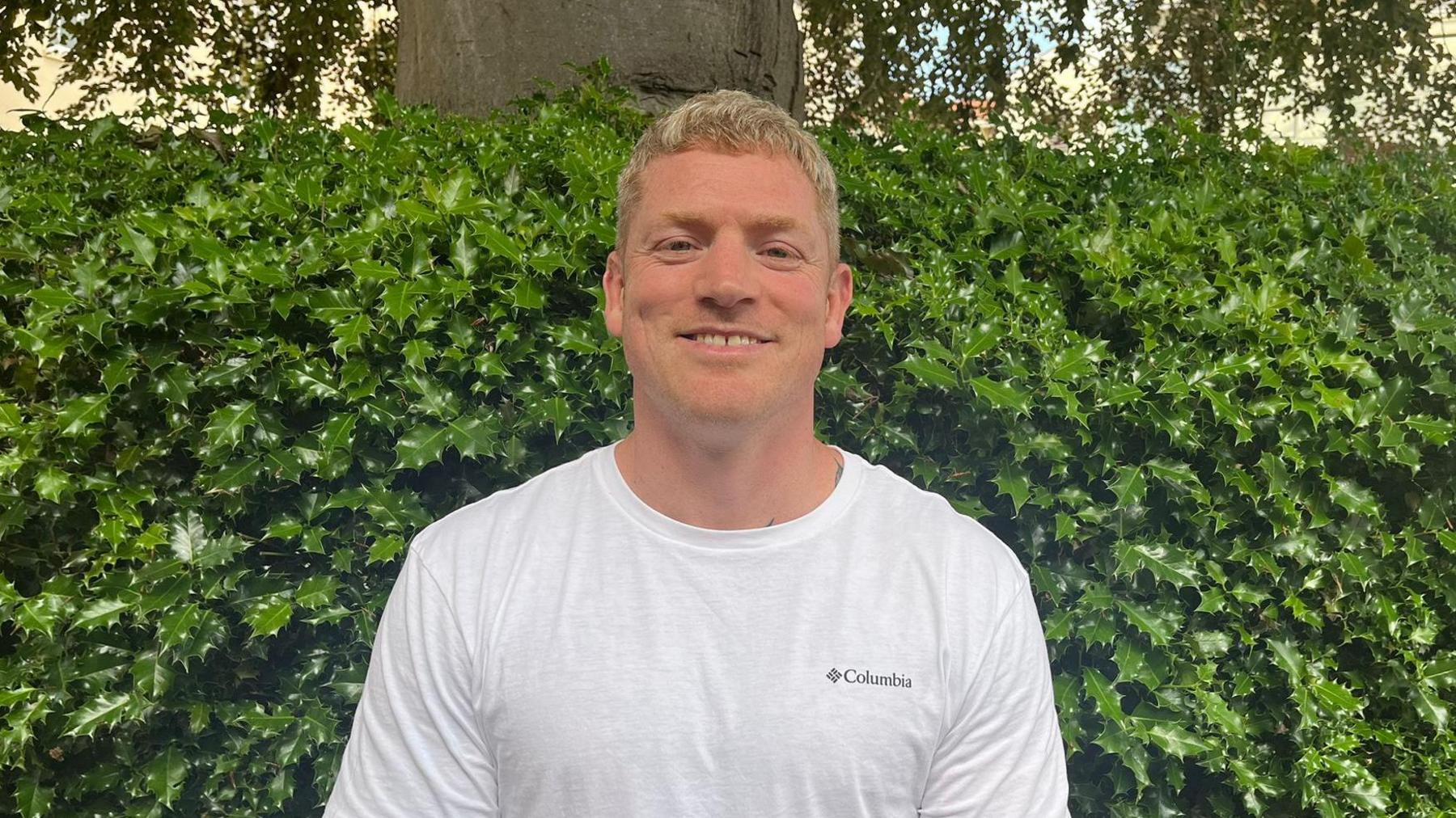 Jason standing in front of a tree and bush wearing a white T-shirt and smiling at the camera