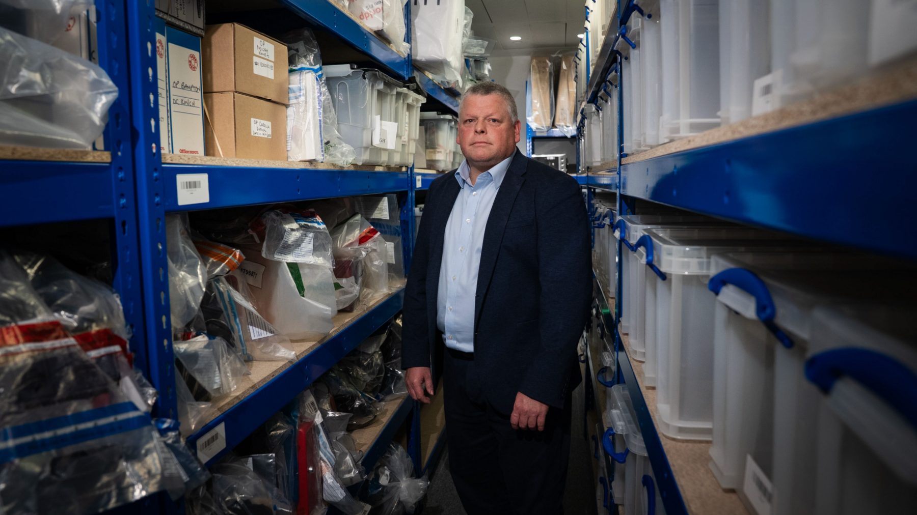 Paul Curtis stands facing the camera in the aisle of the evidence room, piled high with luxury goods seized from financial fraudsters
