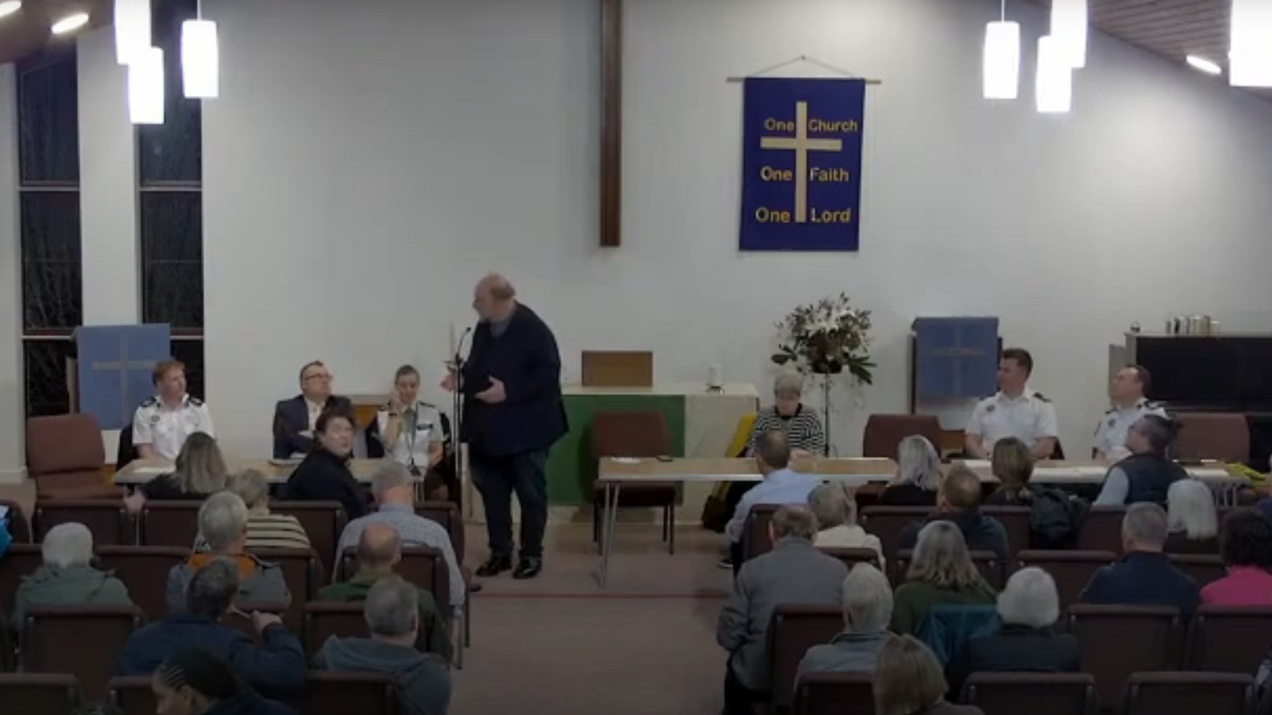 A screenshot of a live feed from inside St Mark's where the pews are full of residents, seen from behind, listening to Tom Wootton who is talking into a microphone at the front. Officers from the police and fire service are sat on desks facing the audience.