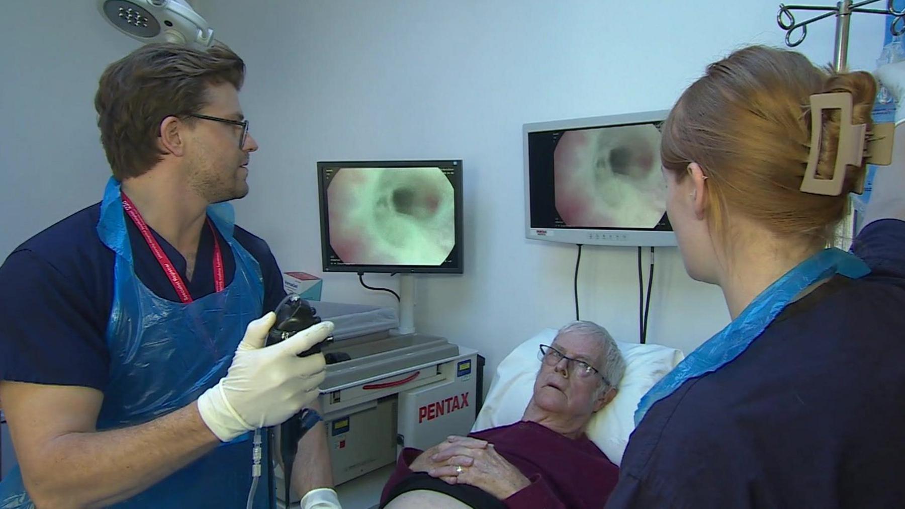 Two healthcare workers in blue scrubs, wearing gloves and plastic aprons. One of them is holding a remote control. In the middle of them is a man on a hospital bed. Two monitor screens above their heads show the inside of a urethra