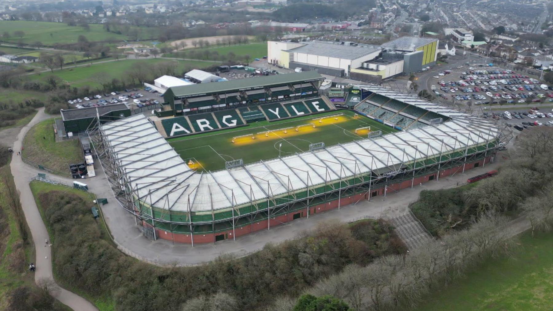 A drone shot of Home Park Stadium, home of Plymouth Argyle Football Club. It's a grey day with the drone looking across the pitch and stands. The ground is surrounded by trees with a large building and car park to the right of the image.