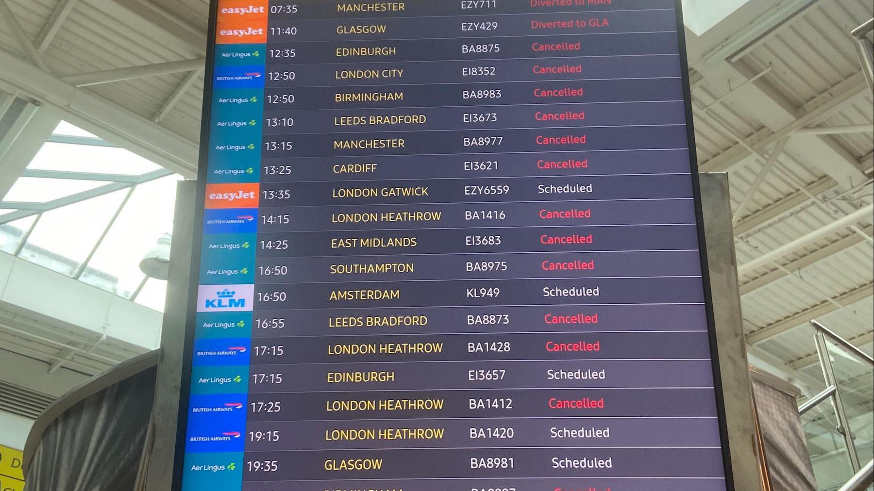 A flight arrivals board at Belfast City Airport. Most have the tag, in red font, 'Cancelled' beside the destination. In white writing some say 'Scheduled.'