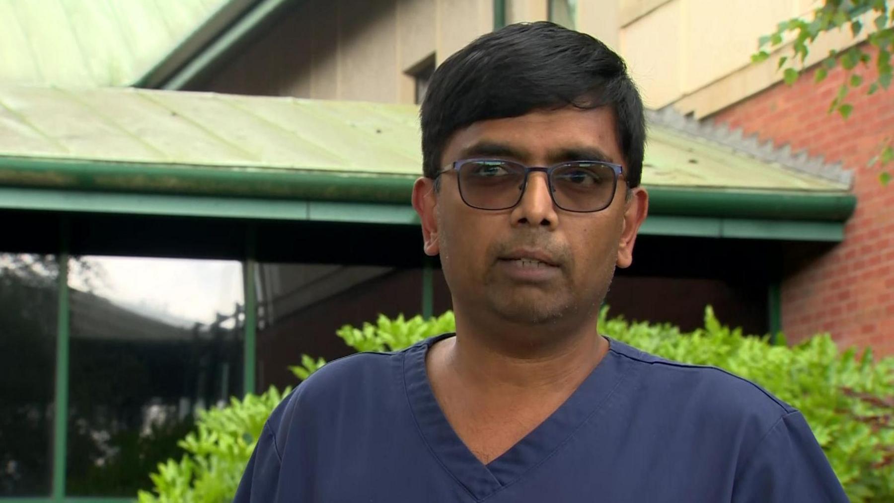 An Asian man standing outside wearing medical scrubs. A building and some shrubbery in the background