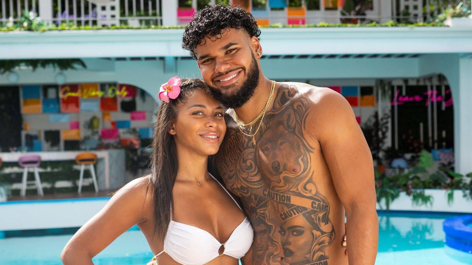 Amaya Espinal and Bryan Arenales pictured in the Love Island USA villa. Amaya has her brown hair pinned back with a pink flower and poses with her hand on her hip while wearing a white bikini. Bryan leans into Amaya smiling. He has curly dark hair and a short beard. He is topless, revealing a heavily tattooed chest with images of wolves, women and caution tape. They're pictured in front of a swimming pool and bar.