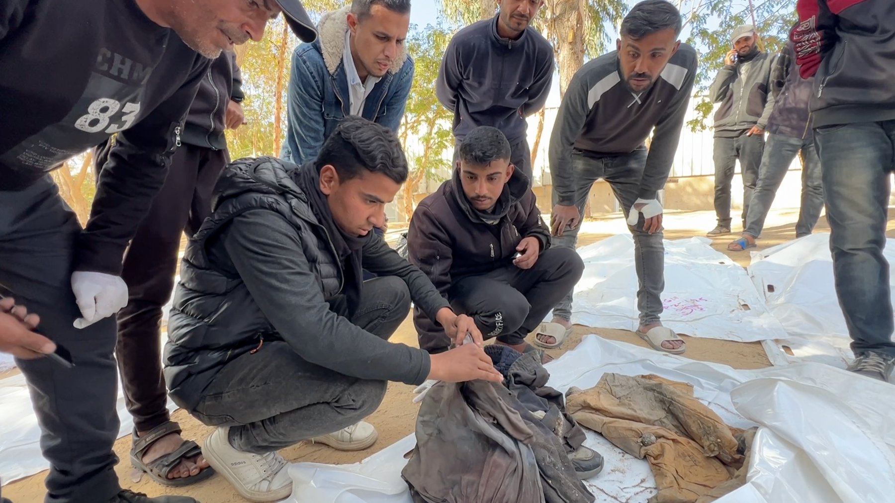 Young men crouch over clothes in a white body bag