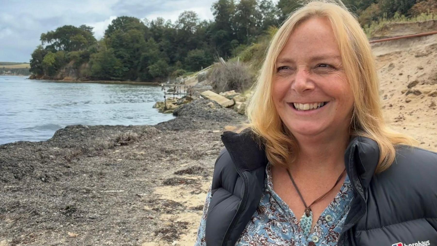 Tracey Church stands on a section of sandy beach, with some water and a green forest of trees in the background. She is smiling and looks slightly off camera. Tracey has shoulder length strawberry blonde hair styled in a side parting. She has a blue buttoned blouse with a brown and white paisley print on it and wears a blue stone necklace on a piece of leather around her neck. She has a slate grey puffer gilet over the top of her blouse.