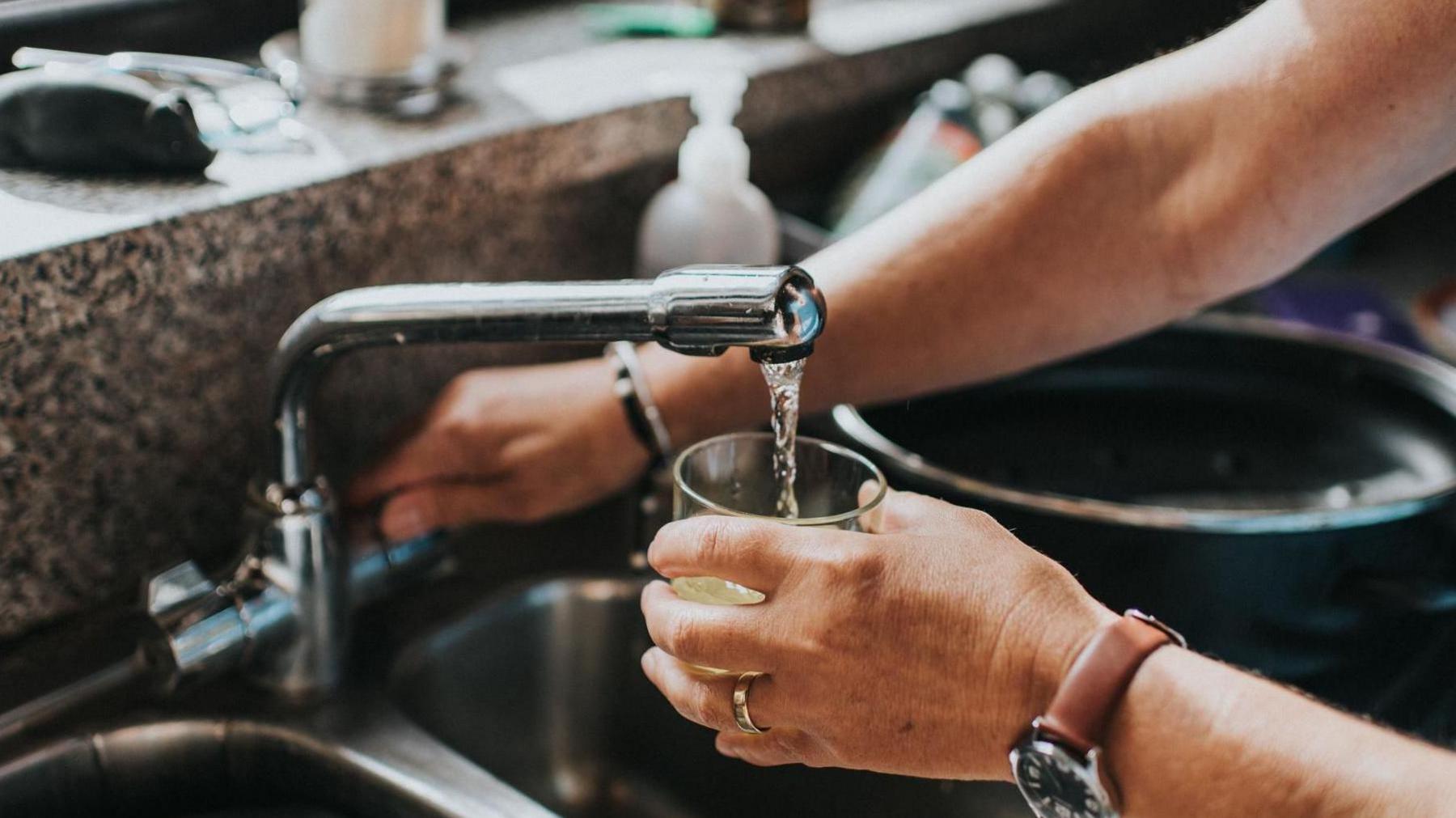 A person pouring water into a glass from a tap. One hand is holding the glass whilst the other turns the tap. In the background the rest of the silver sink, a black pan and the stone windowsill is seen.