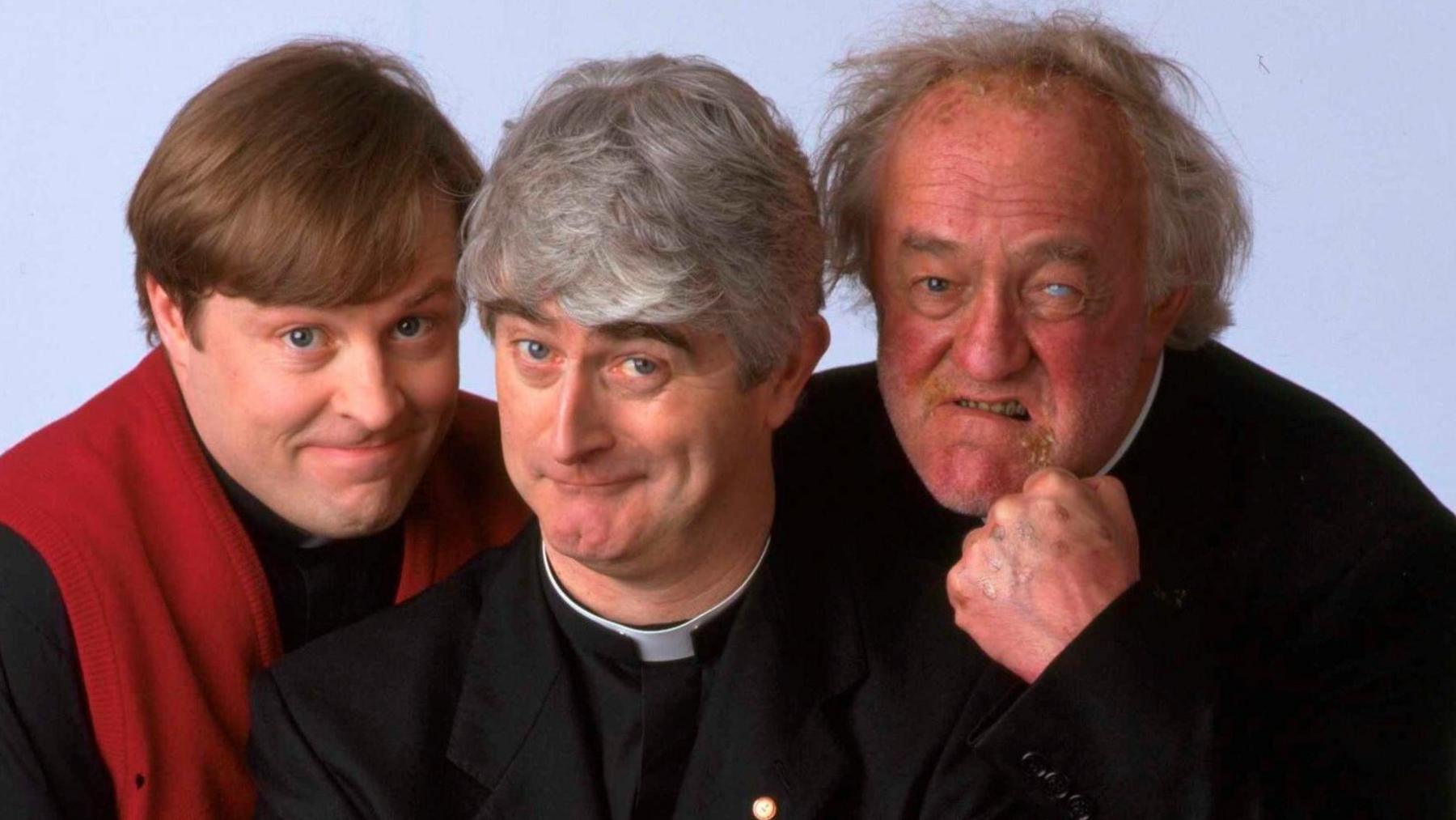 The three actors who played priests in the Father Ted TV show standing side by side and looking into the camera. They are all dressed as priests. Father Dougal, on the left, is wearing a red jumper. Father Ted in the middle, has grey hair and is looking happy. Father Jack on the right is brandishing a fist and looking aggressive.