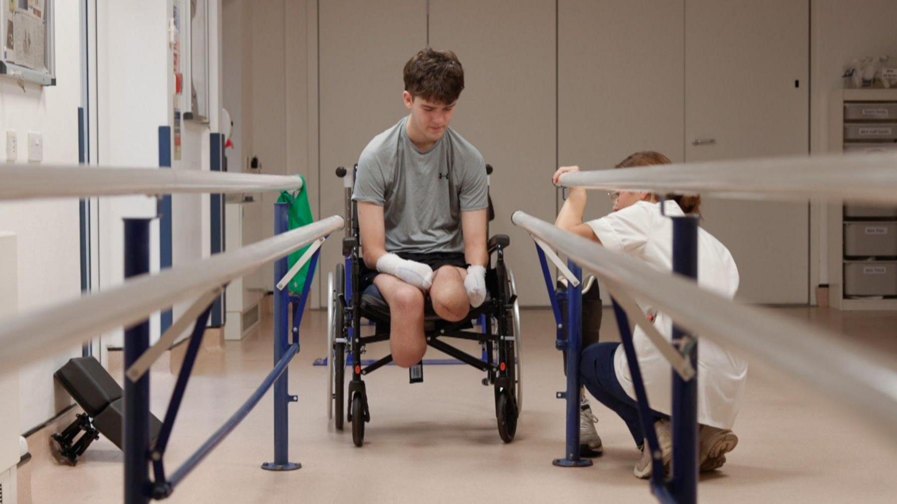 Hamish Wilson sat in a wheelchair before walking for the first time on his new prosthetic legs. There is a nurse crouching down next to him. 