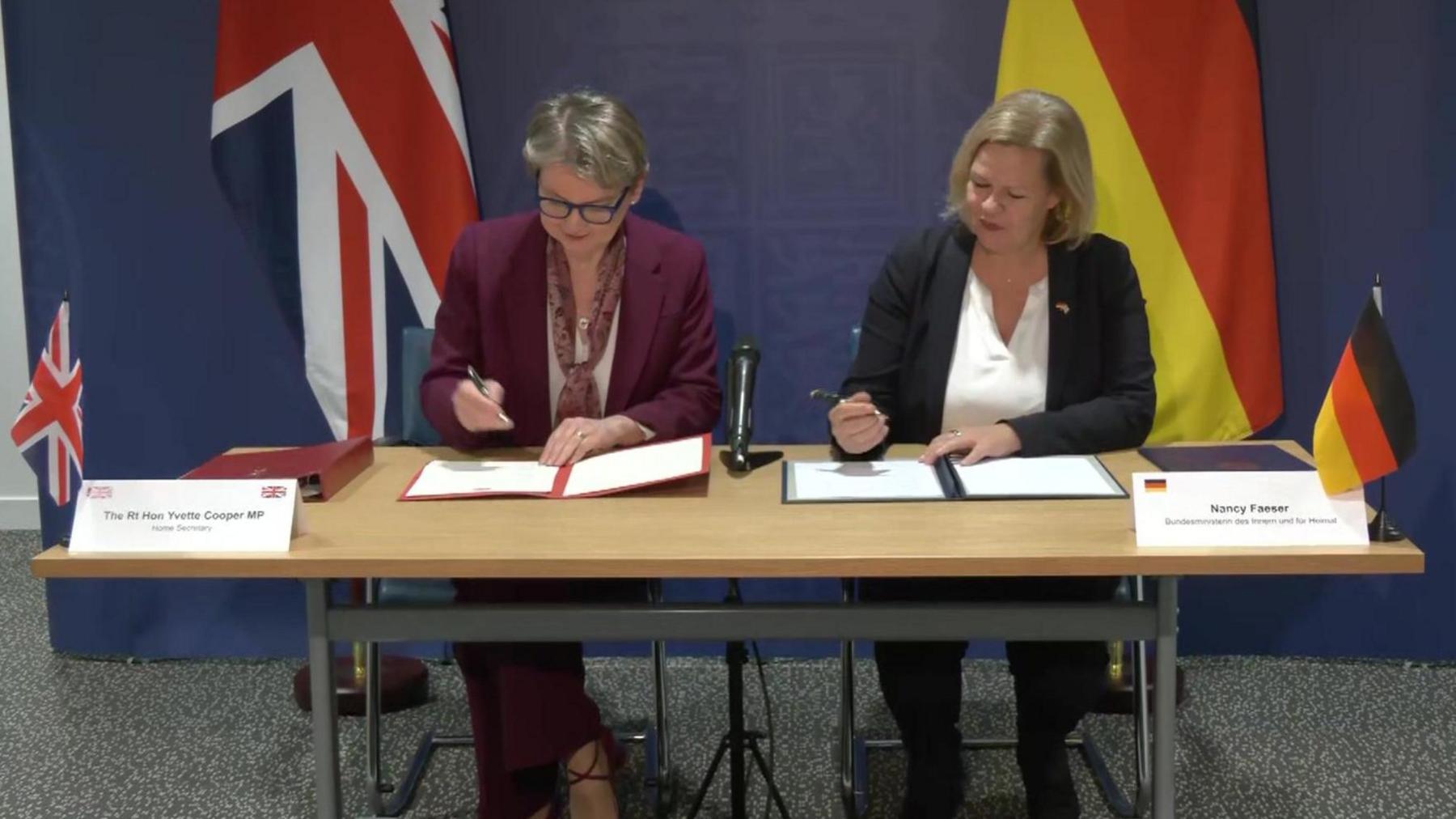 UK Home Secretary Yvette Cooper and German Interior Minister Nancy Faeser sign agreements, while sitting in front of the UK and Germany flags