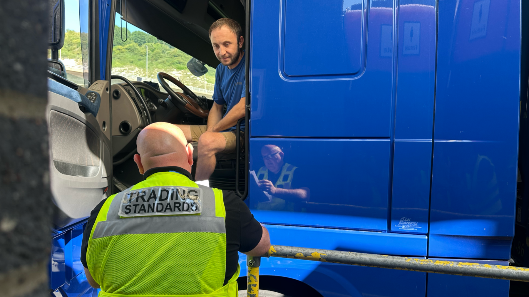 A trading standards officer checking the passport and documents of a lorry driver, who has his door open and is sitting inside the cab of his blue lorry