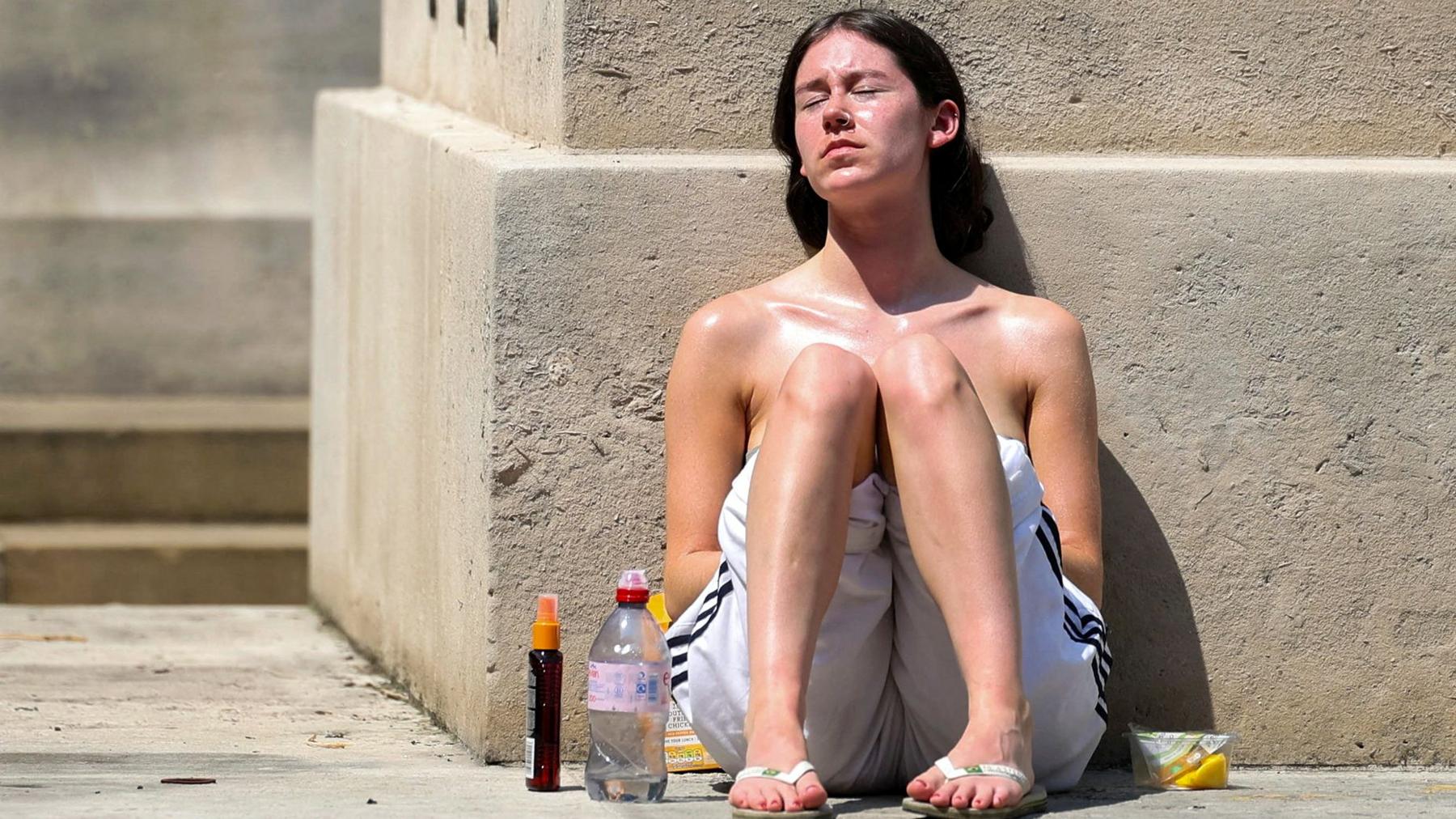 A woman sits on the ground and leaning against a beige stone wall with her knees up near her chest in the blazing sun. She has a bottle of water on the ground