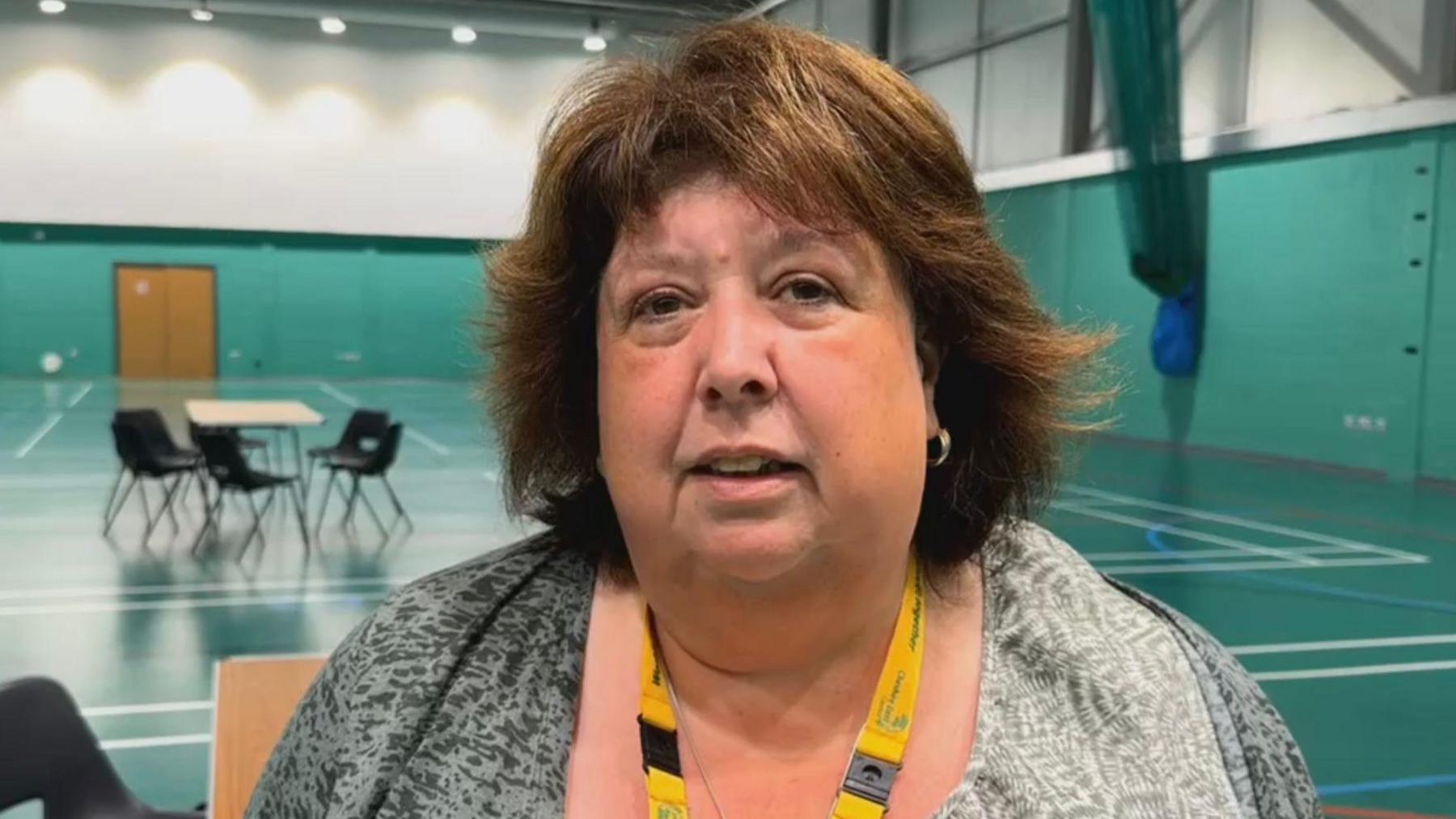 A woman with long brown hair and wearing a light grey top and a yellow lanyard around her neck, stands in the middle of a sports hall with tables and chairs behind her.