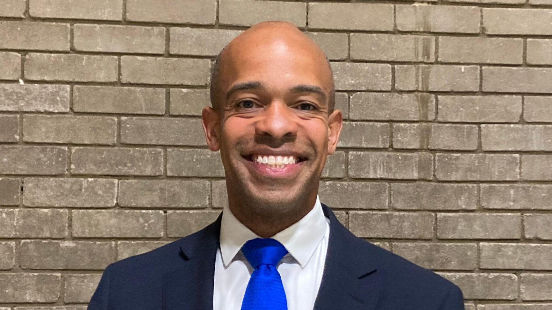 Ben Obese-Jecty is smiling in front of a grey brick wall. He is wearing a dark blazer, white shirt and bright blue tie.