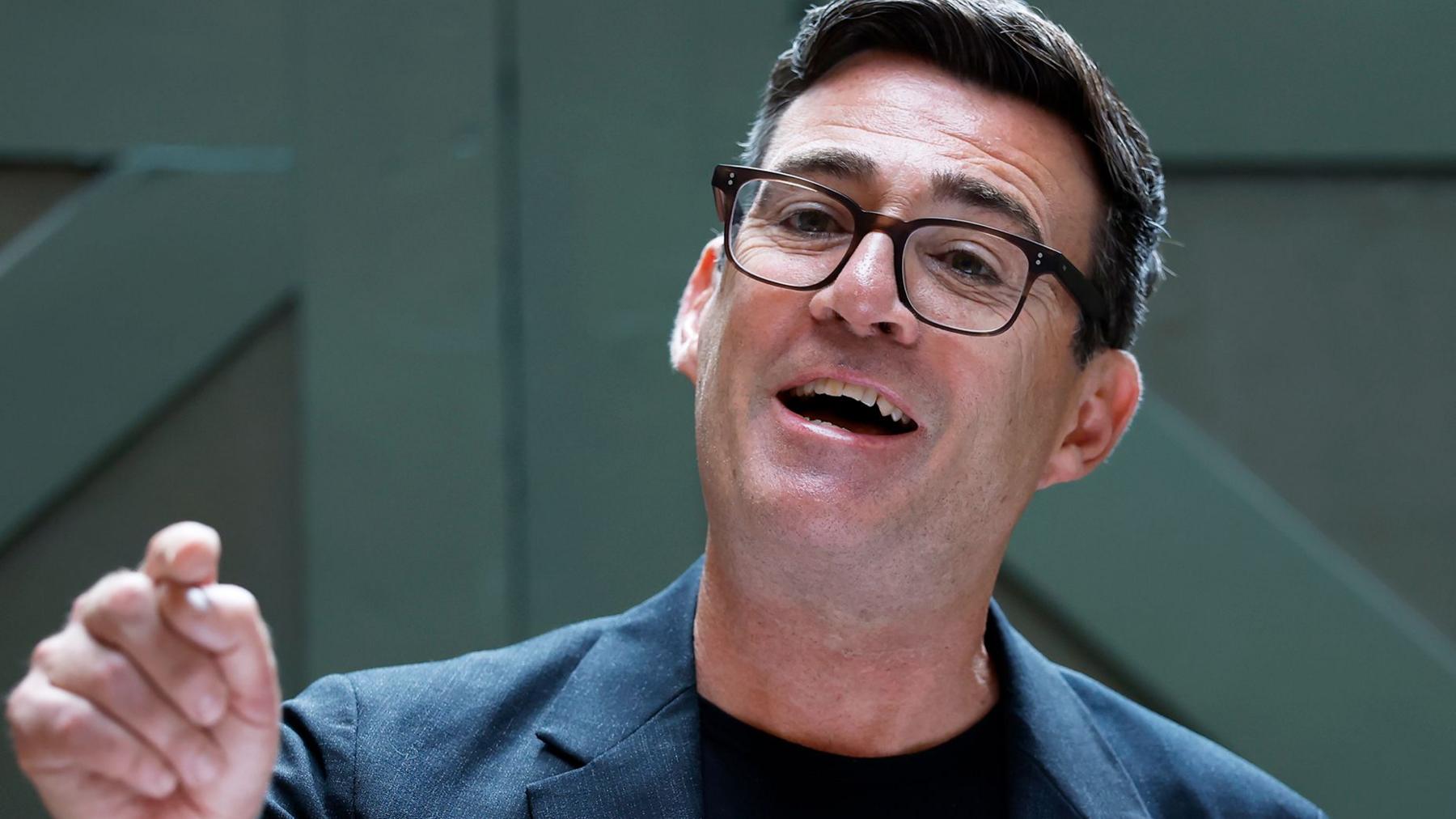 Andy Burnham speaking and smiling in front of a green door wearing a dark suit and glasses