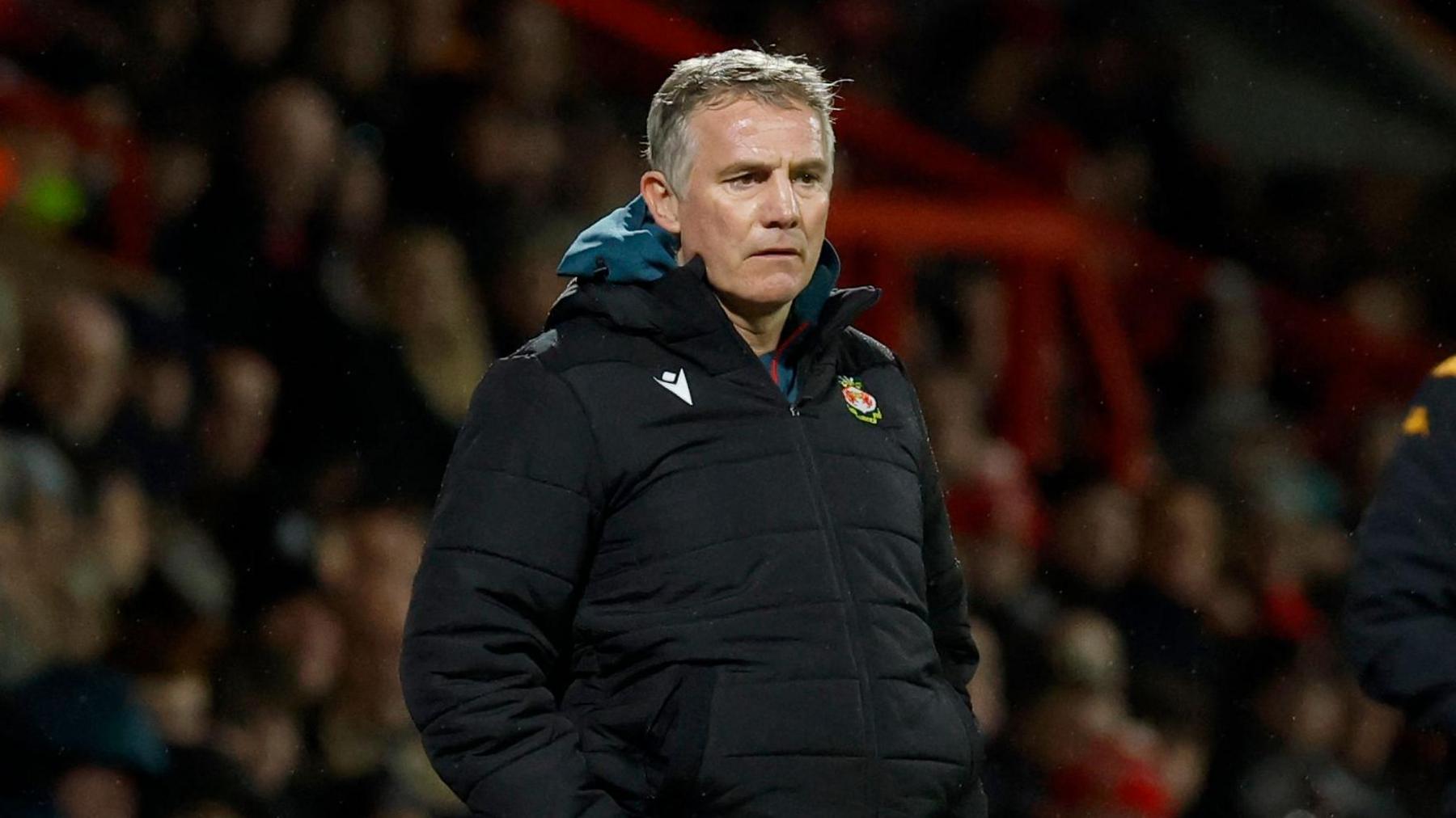 Wrexham manager Phil Parkinson wearing a black puffer coat with the Wrexham badge on his chest and his hands in his coat pockets.