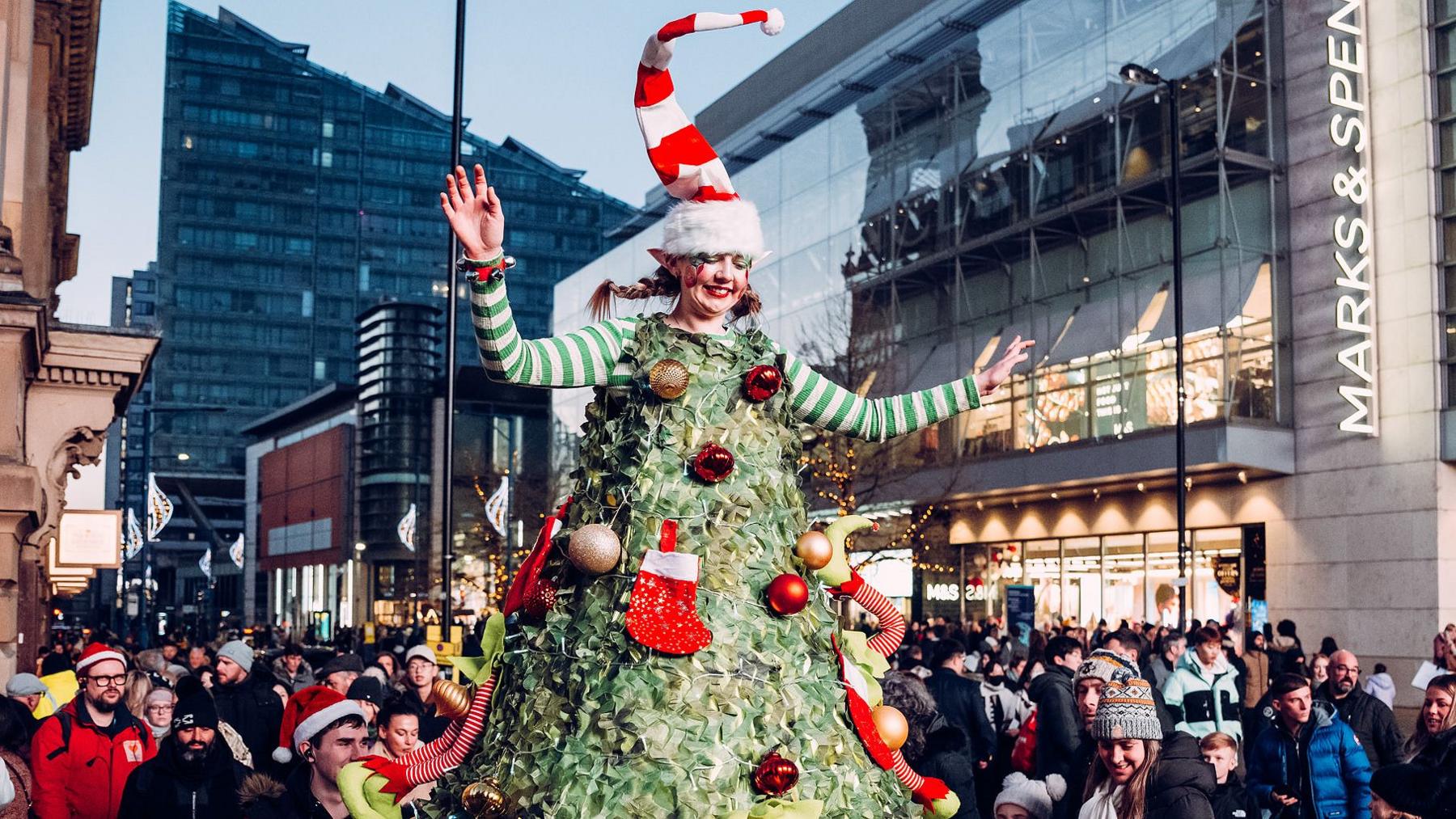 A woman dressed up as a Christmas tree on stilts for a Christmas Parade in a city centre with hundreds of people lining the streets.