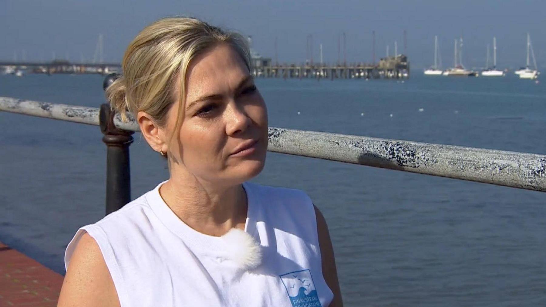 Bianca Carr is looking slightly off camera giving an interview. She is on a bridge, with a metal rail behind her and in the background is a harbour, with the sea and a collection of white boats seen in the distance. Bianca has blonde hair, which is tied back in a low bun and a shorter piece of hair frames her faces. She wears a sleeveless white tank top, which has a blue square Final Straw Foundation logo on it.