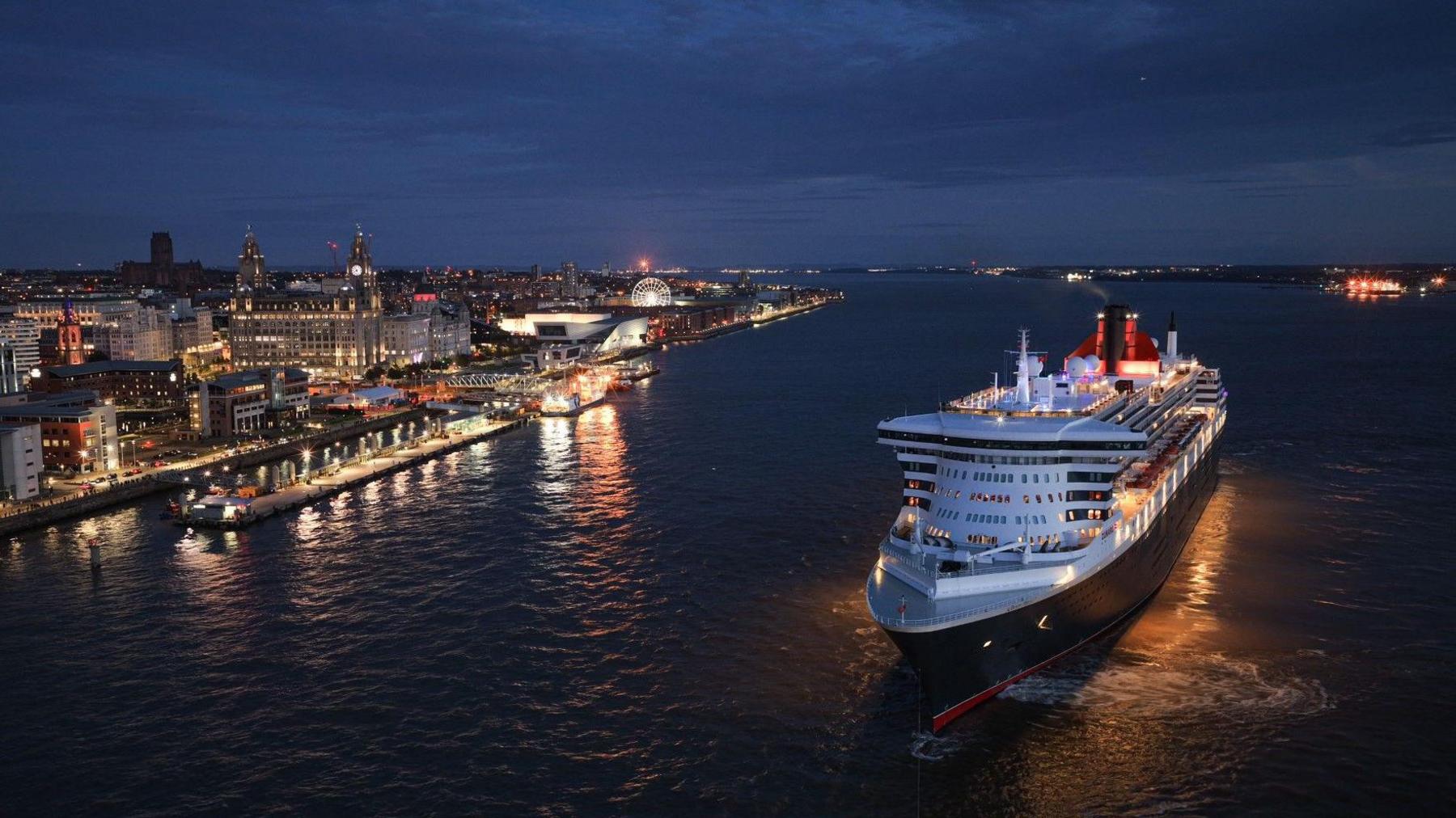 The Queen Mary 2 ocean liner on the River Mersey at night. The Liverpool waterfront and Liver Building is lit up in the background. Lights also line the Wirral waterfront.