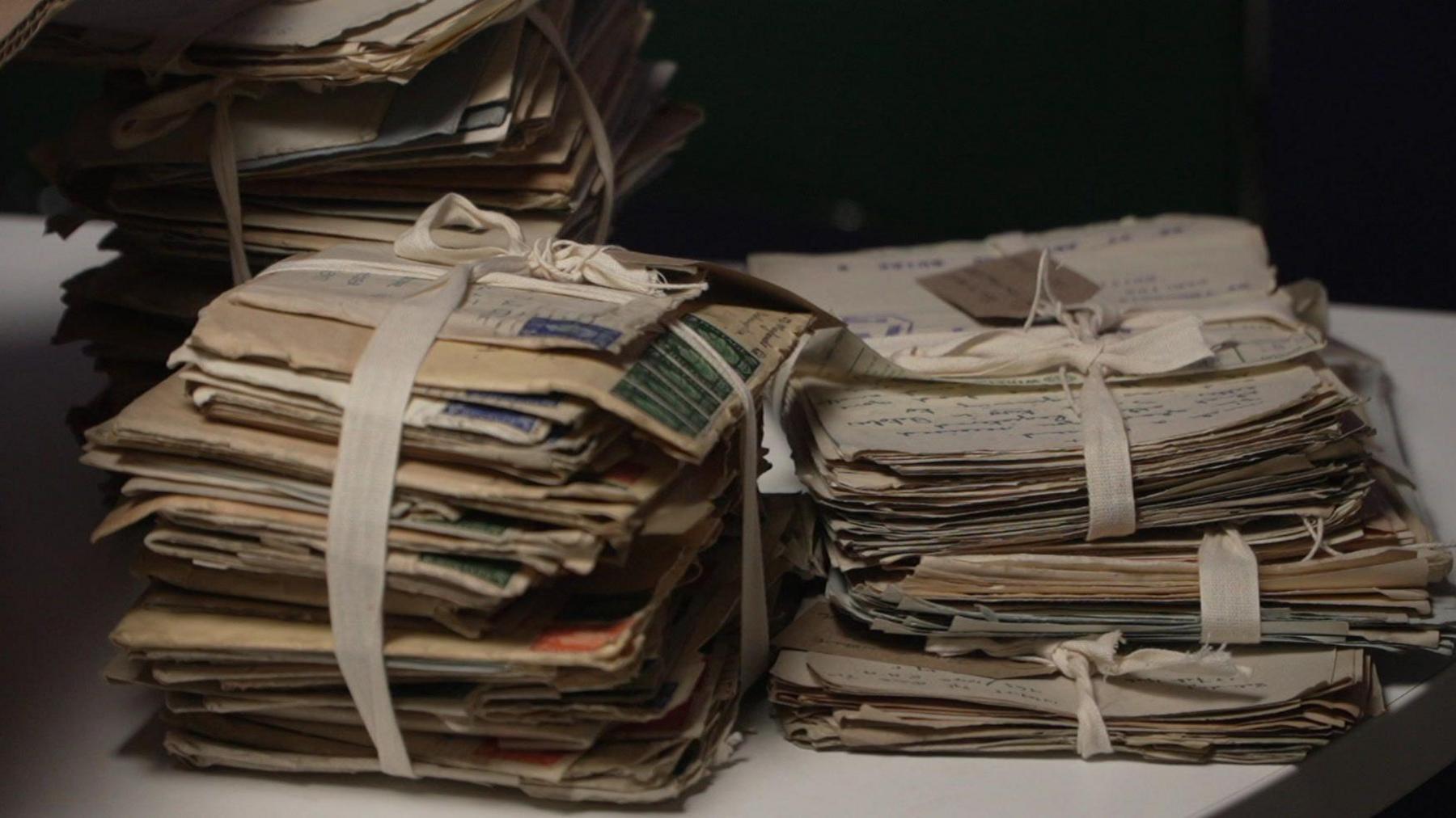Three stacks of hand-written letters resting on a table. They are held in their piles by white bows, which are tied around them.
