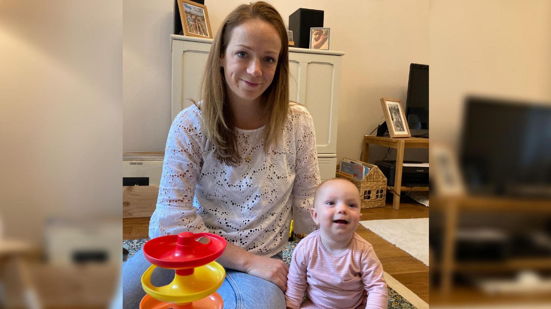 Alice Cook wearing a white blouse and sitting on her living room floor beside her baby, Henrietta, who is wearing a pink stripey baby grow and smiling at the camera. 