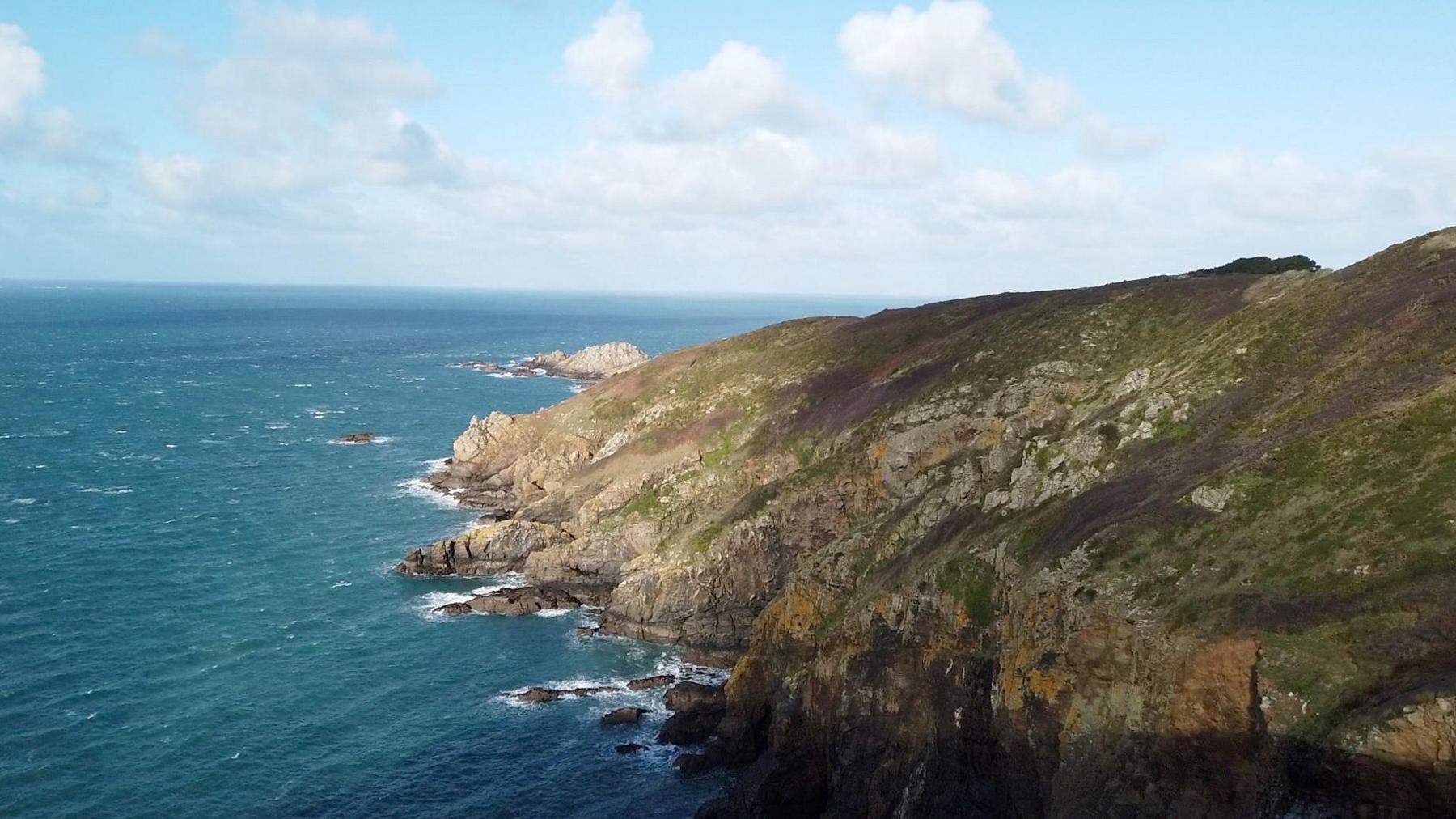 The rocky coastline of Sark with sea and a steep cliff dotted with greenery.
